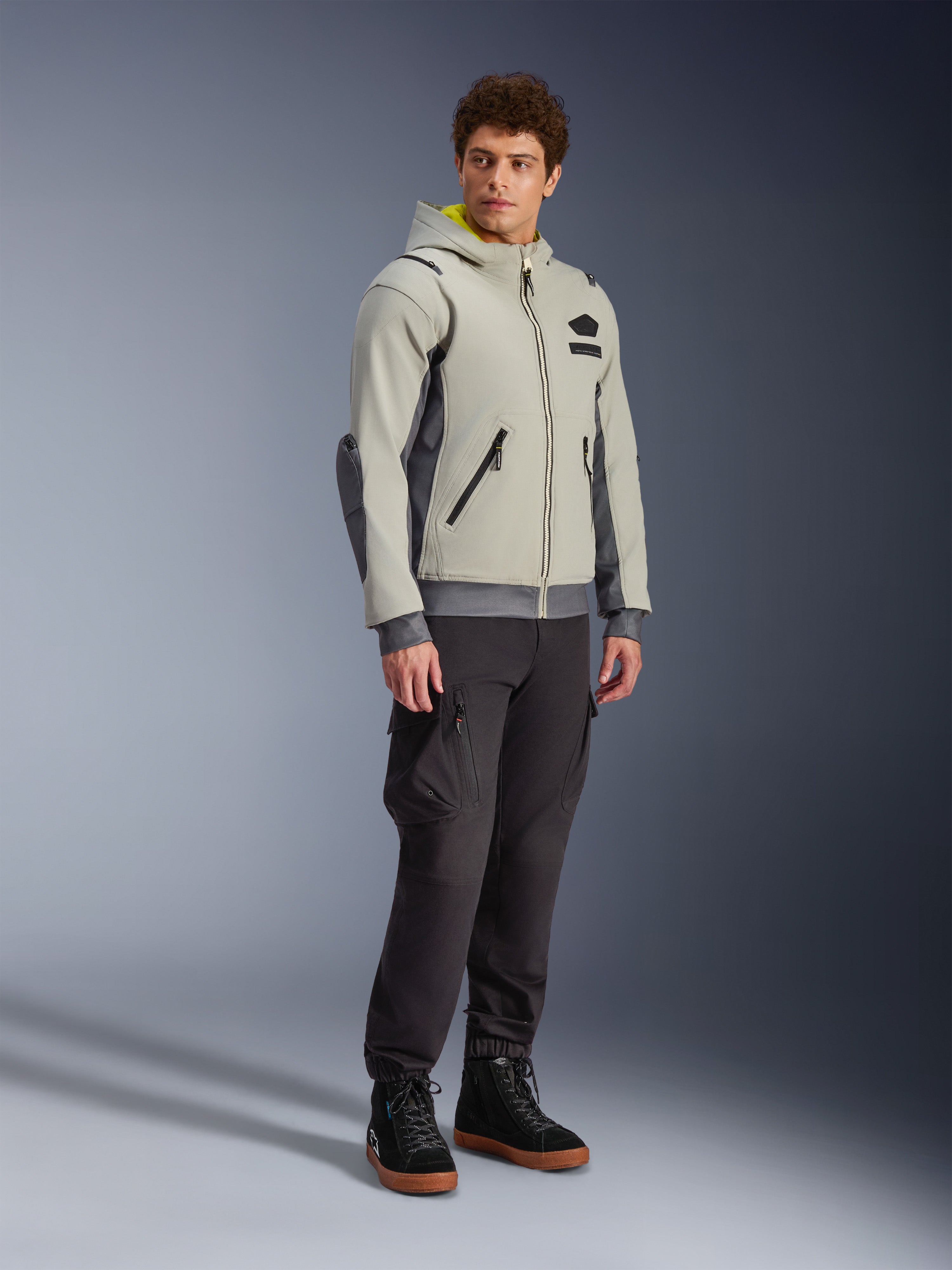 Alpinestars Moblast vandtæt jakke, Iron Gray og Dark Sage, urban motorcykeljakke med hættetrøjedesign, båret af en model med mørke kørebukser og sorte støvler, minimalistisk cyber-tech-æstetik med gult indvendigt for i hætten og vandtætte lynlåse