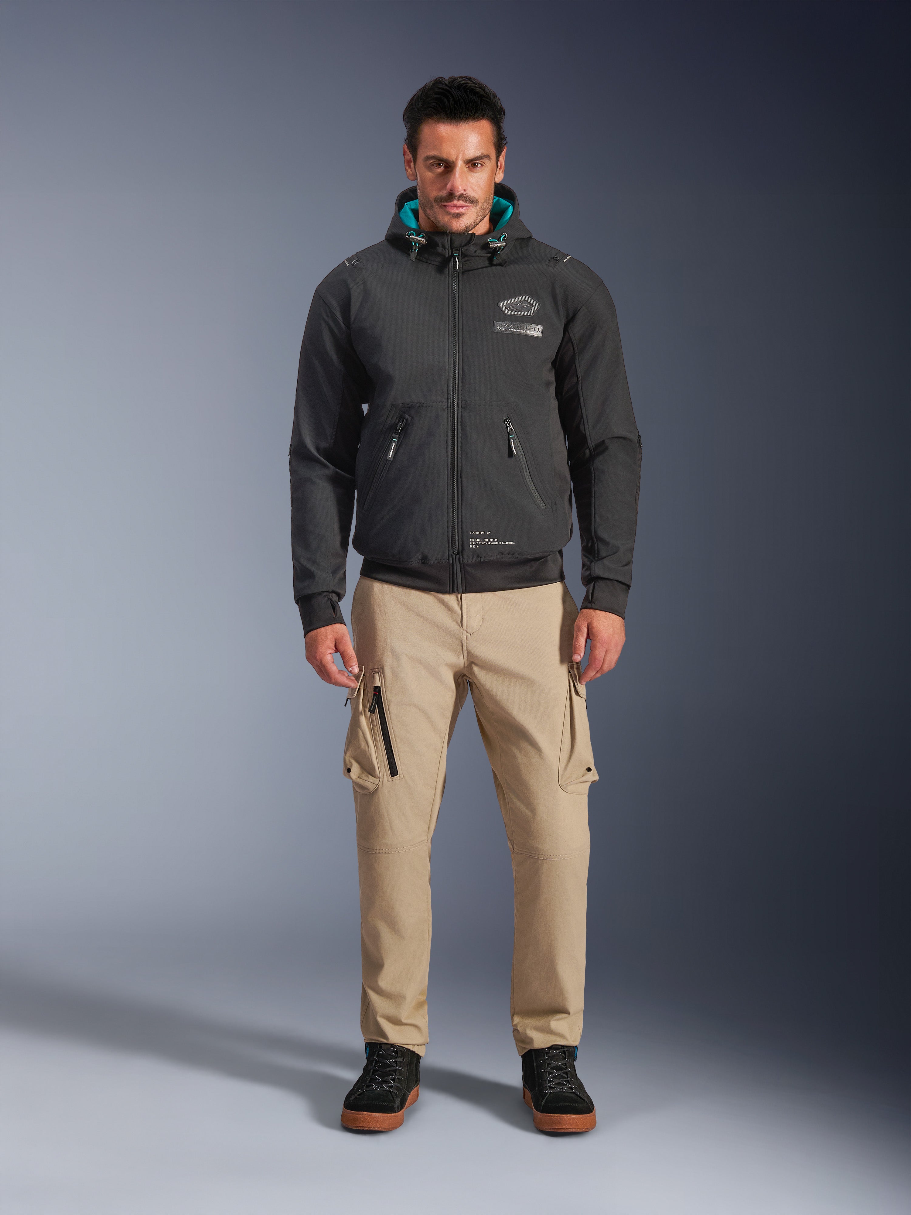 Alpinestars Moblast Waterproof Jacket, sort urban motorcykel-hættetrøje med blågrønt indvendigt hættefor, båret med sandfarvede cargobukser og sorte high-top støvler, moderne køreudstyr til alle årstider