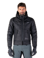 Alpinestars Mohobbs vandtæt jakke, sort polstret bomber-motorcykeljakke, quiltet poly-materiale med lynlås foran og lynlåslommer, lille hvidt logo på brystet, båret af en model med grå bukser og handsker, urban streetwear-design til bykørsel