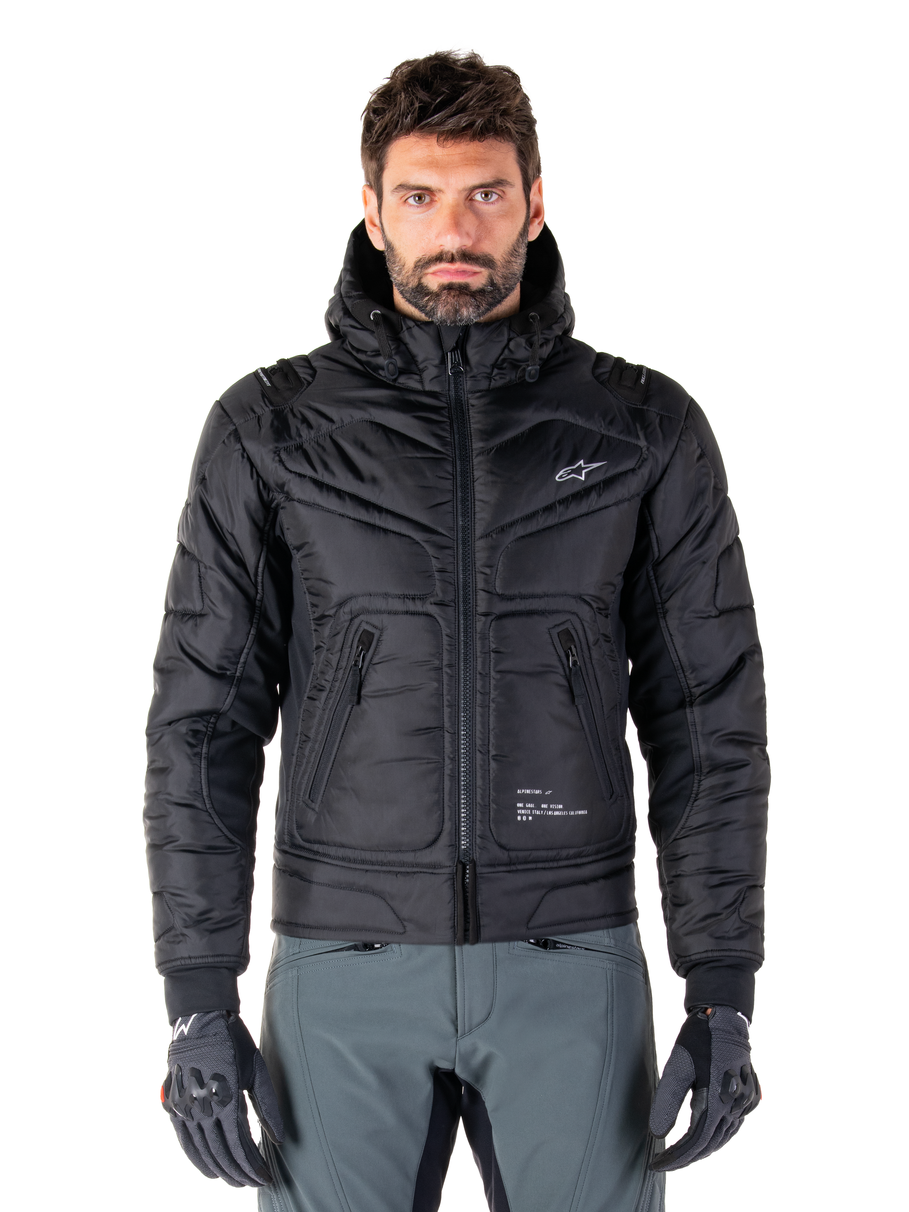 Alpinestars Mohobbs vandtæt jakke, sort polstret bomber-motorcykeljakke, quiltet poly-materiale med lynlås foran og lynlåslommer, lille hvidt logo på brystet, båret af en model med grå bukser og handsker, urban streetwear-design til bykørsel