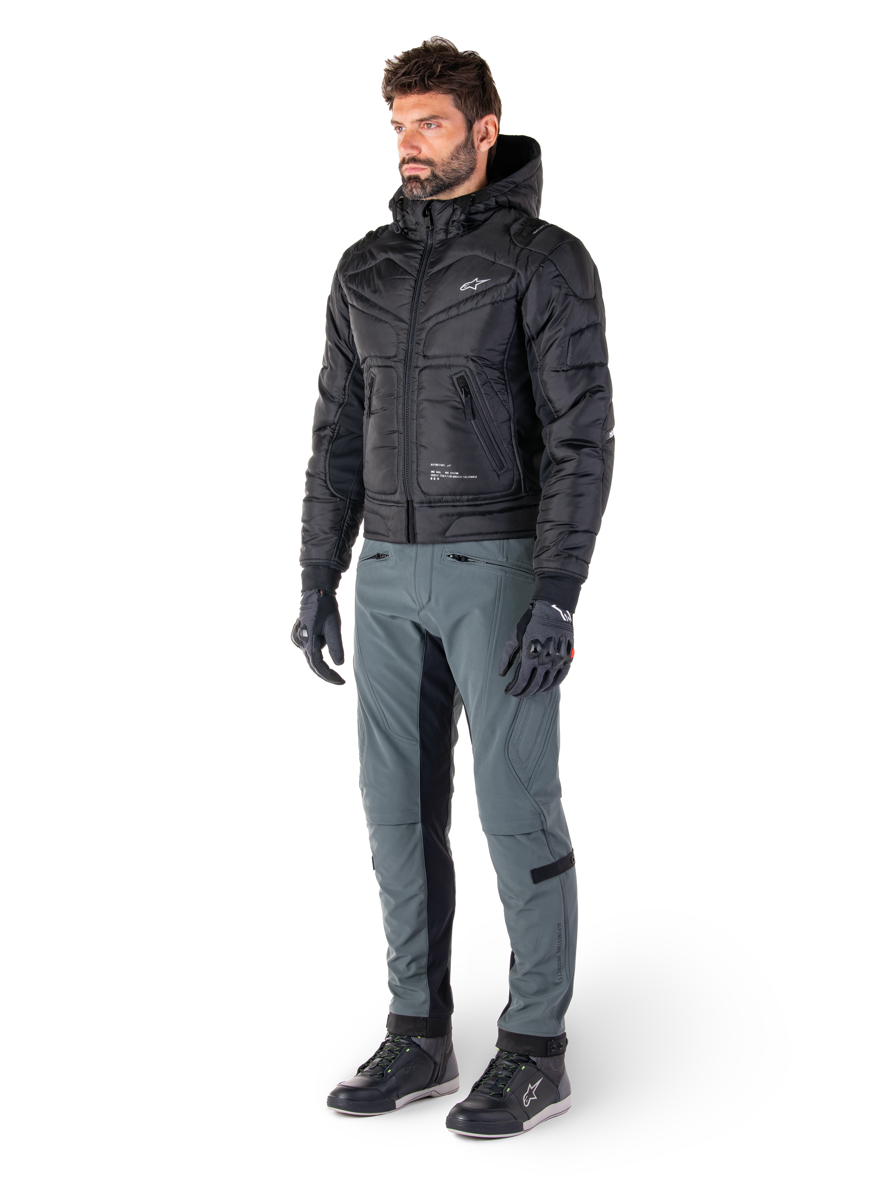 Alpinestars Mohobbs Waterproof Jacket, sort, quiltet urban motorcykel-bomberjakke med hætte, båret af en model med grå kørebukser og sorte handsker, med hvidt brystlogo, moderne streetwear-inspireret beskyttelsesudstyr