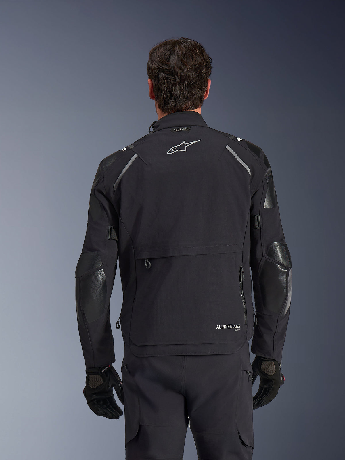Alpinestars Andes Pro Drystar® XF-jakke, sort og mørkegrå, set bagfra båret af en mandlig model, med et stort opbevaringsrum bagpå, reflekterende detaljer på skuldrene og logo-branding, kombineret med sorte handsker og bukser til adventure touring.
