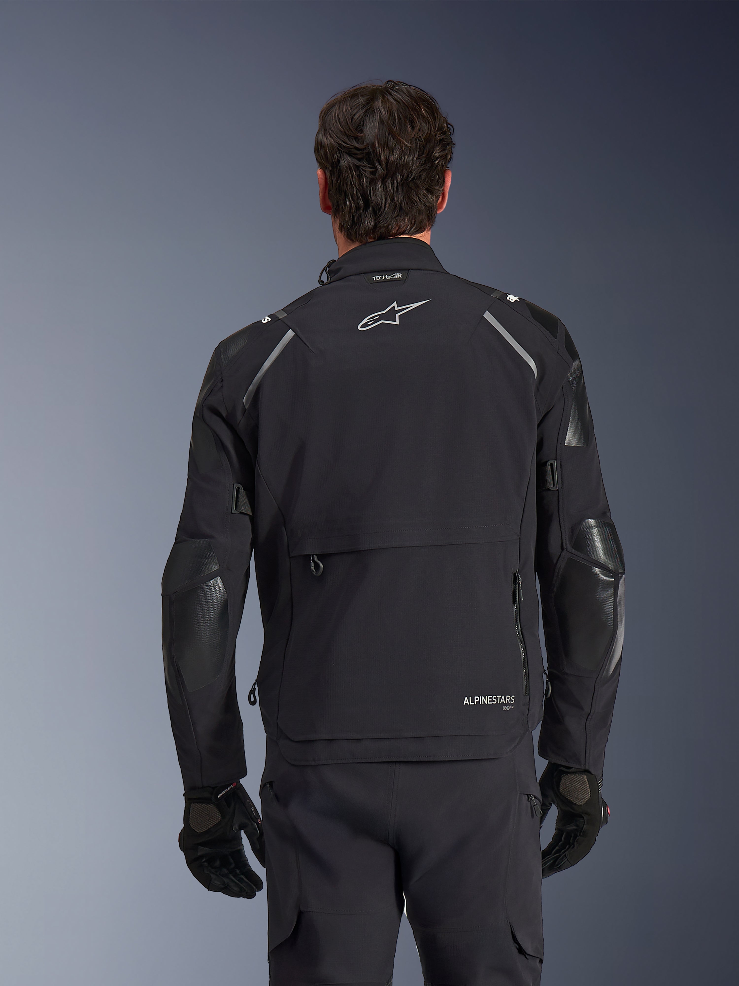 Alpinestars Andes Pro Drystar® XF-jakke, sort og mørkegrå, set bagfra båret af en mandlig model, med et stort opbevaringsrum bagpå, reflekterende detaljer på skuldrene og logo-branding, kombineret med sorte handsker og bukser til adventure touring.
