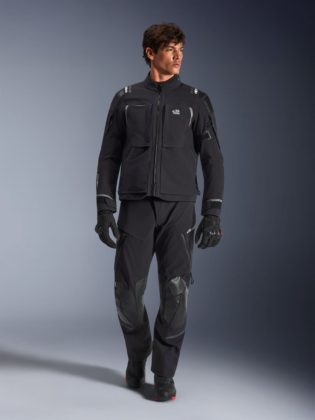 Mandlig model iført Alpinestars Andes Pro Drystar® XF jakke, sort og mørkegrå adventure-motorcykeljakke, kombineret med matchende tekniske bukser og handsker, med flere lommer og 'Terra'-branding på brystet.