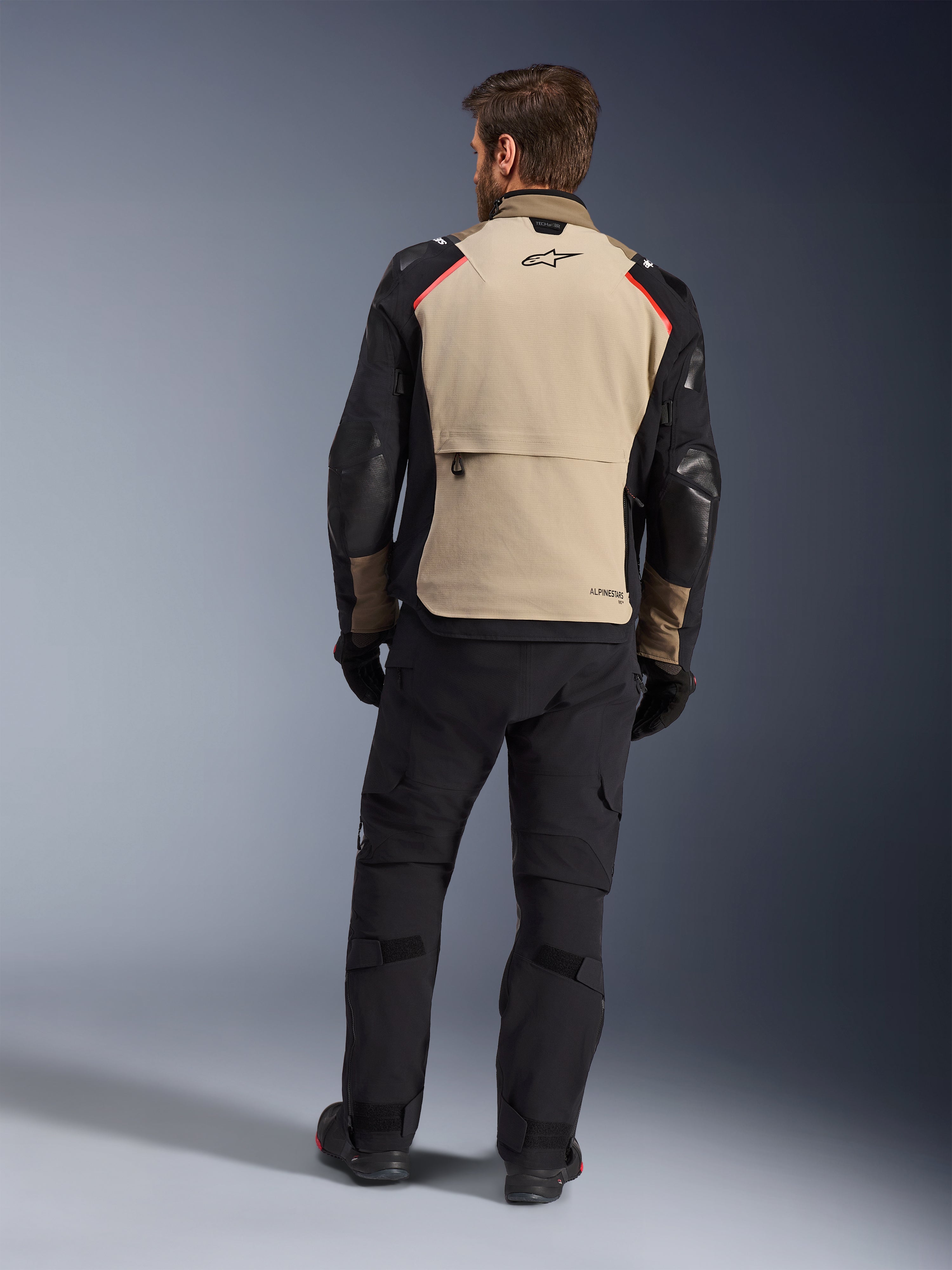 Alpinestars Andes Pro Drystar® XF jakke, Walnut Aluminum, set bagfra, motorcykel adventure-jakke med sorte ærmer og røde skulderdetaljer, båret med sorte kørebukser og handsker.