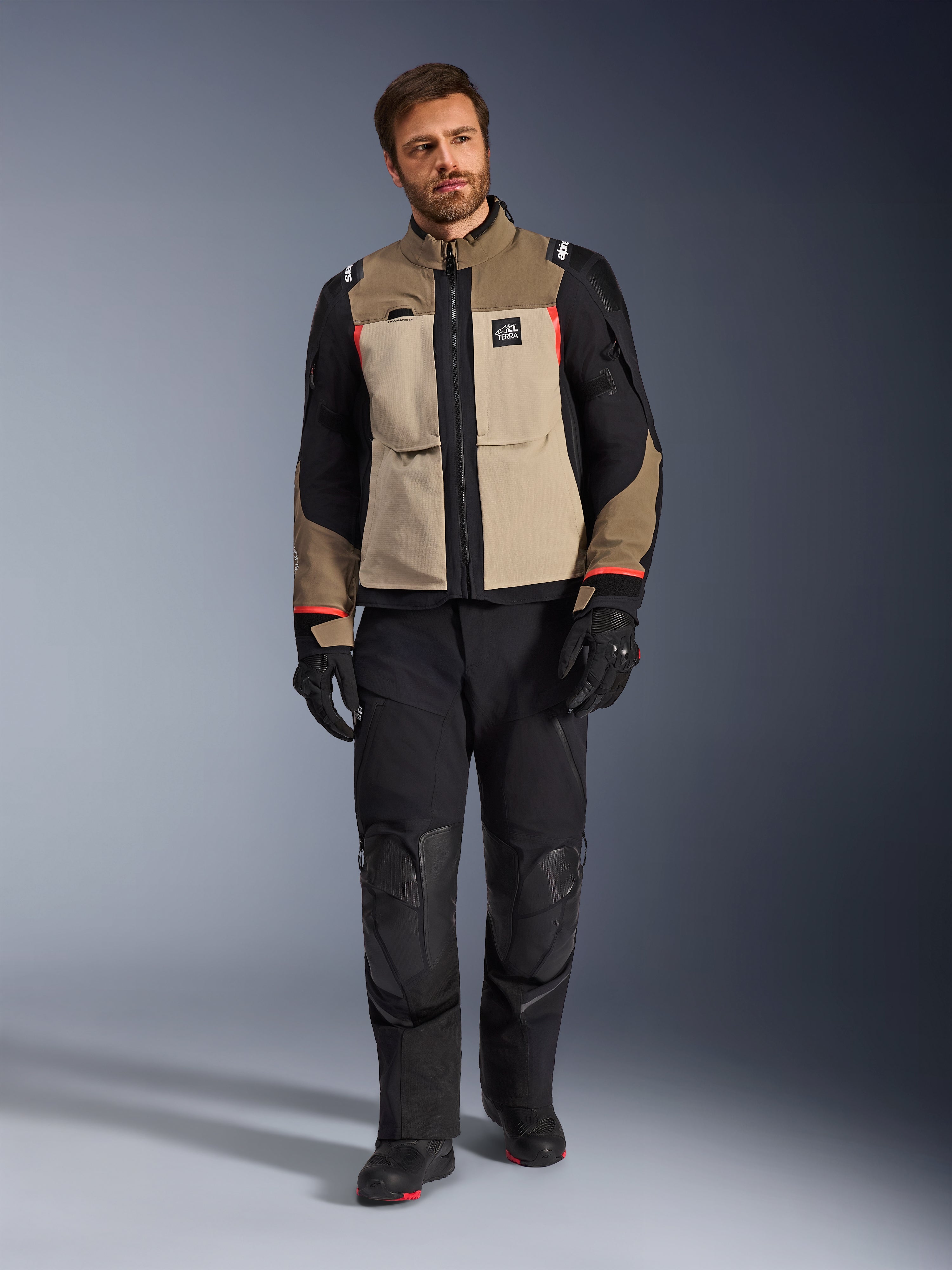Mandlig model iført Alpinestars Andes Pro Drystar® XF-jakke i Walnut Aluminum, tan- og sort adventure-touringjakke med røde detaljer, kombineret med sorte motorcykelbukser, handsker og støvler, studiefoto i fuld figur af beskyttende køreudstyr.