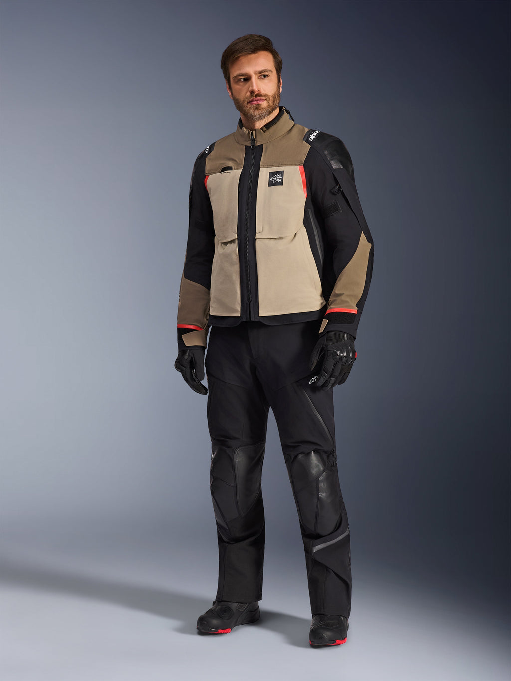 Mandlig model iført Alpinestars Andes Pro Drystar® XF-jakke i Walnut Aluminum, kombineret med sorte motorcykelbukser, handsker og støvler, med et tan og sort multi-panel design med røde detaljer til adventure touring.