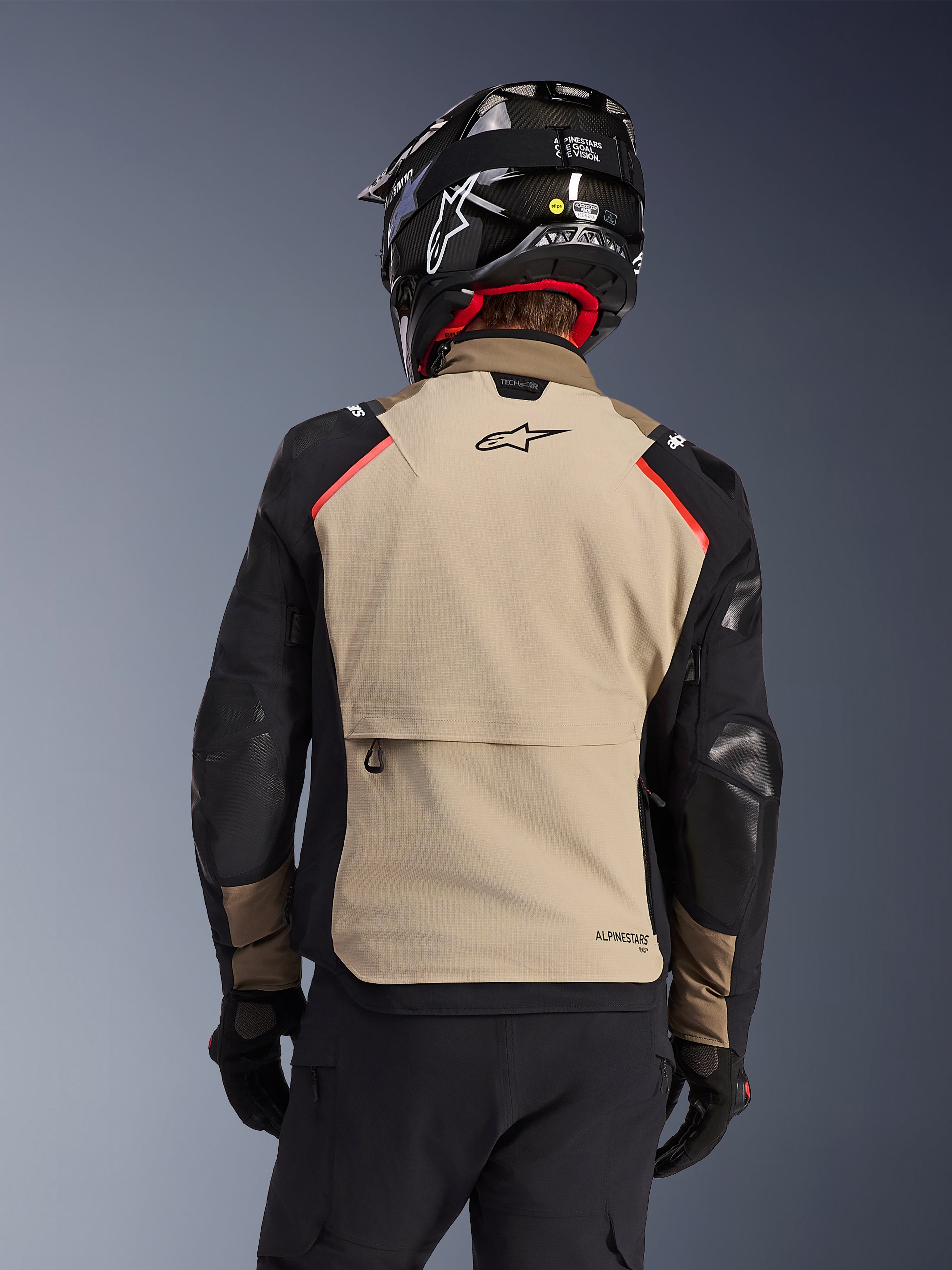 Alpinestars Andes Pro Drystar® XF jakke i Walnut Aluminum, set bagfra, sandfarvet rygpanel med stor cargolomme, sorte ærmer med forstærkede paneler, Alpinestars-logo på den øverste del af ryggen, kombineret med sort hjelm og handsker til adventure touring.