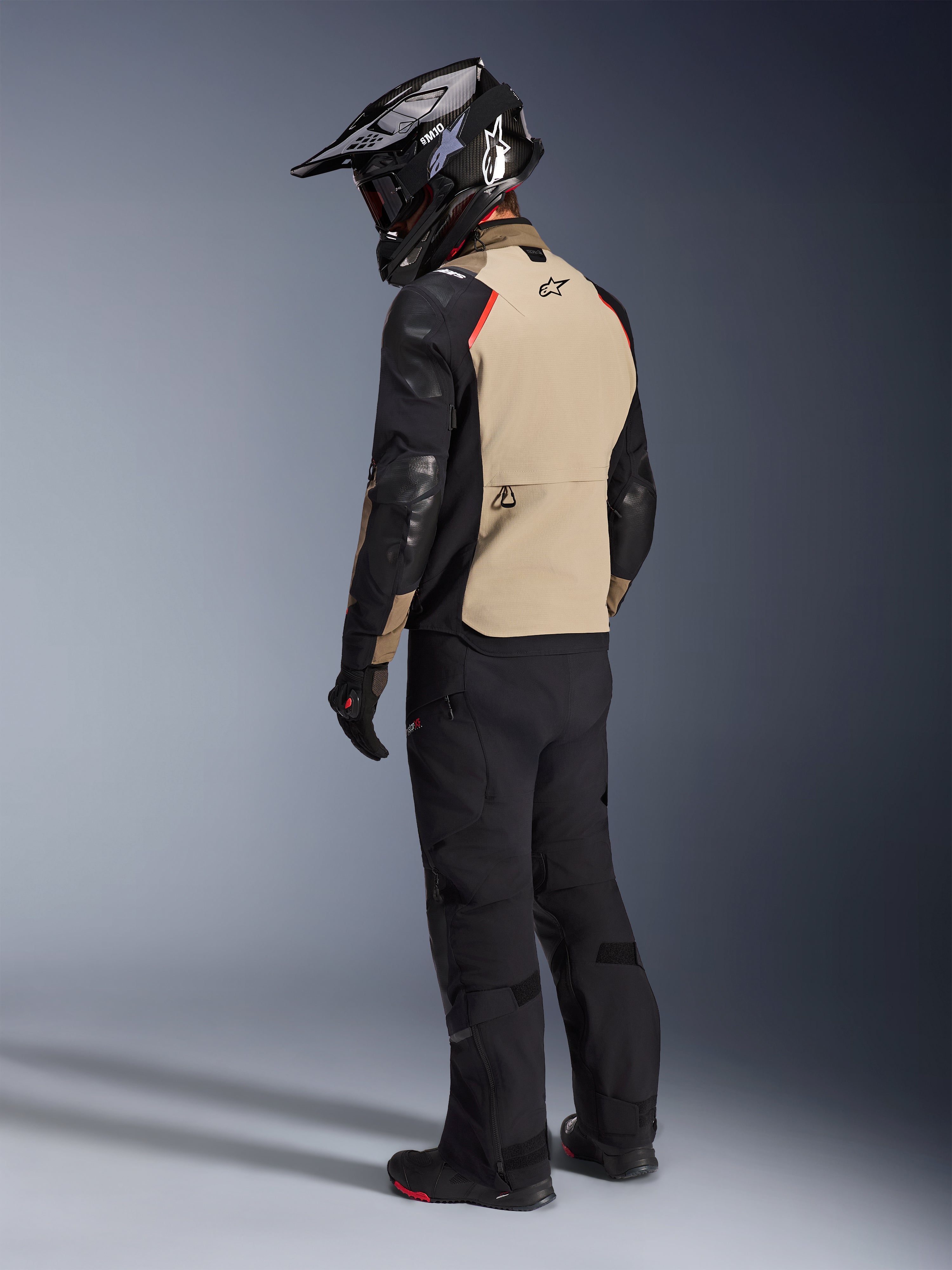 Motorcykelrytter set bagfra iført Alpinestars Andes Pro Drystar® XF-jakken i Walnut Aluminum, med et sandfarvet rygpanel, sorte ærmer og røde detaljer, sort adventure-hjelm, sorte kørebukser og sorte støvler, professionelt adventure-touring-udstyr.