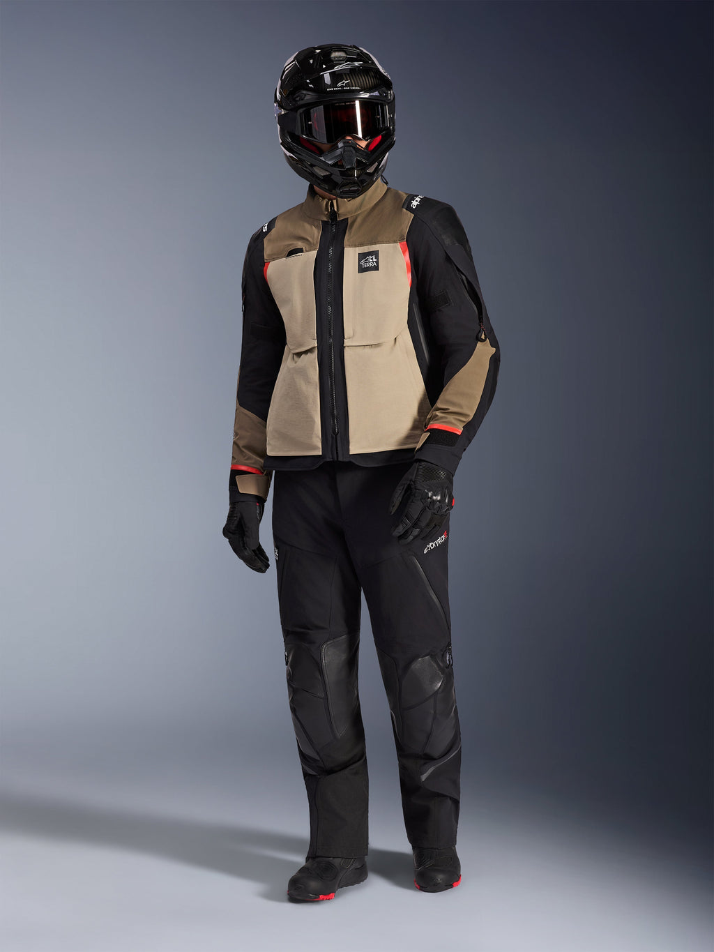 Alpinestars Andes Pro Drystar® XF-jakke i Walnut Aluminum, adventure-motorcykeljakke kombineret med matchende sorte bukser, en sort adventure-hjelm med briller og sorte støvler, med tan-farvede og sorte paneler med røde detaljer.