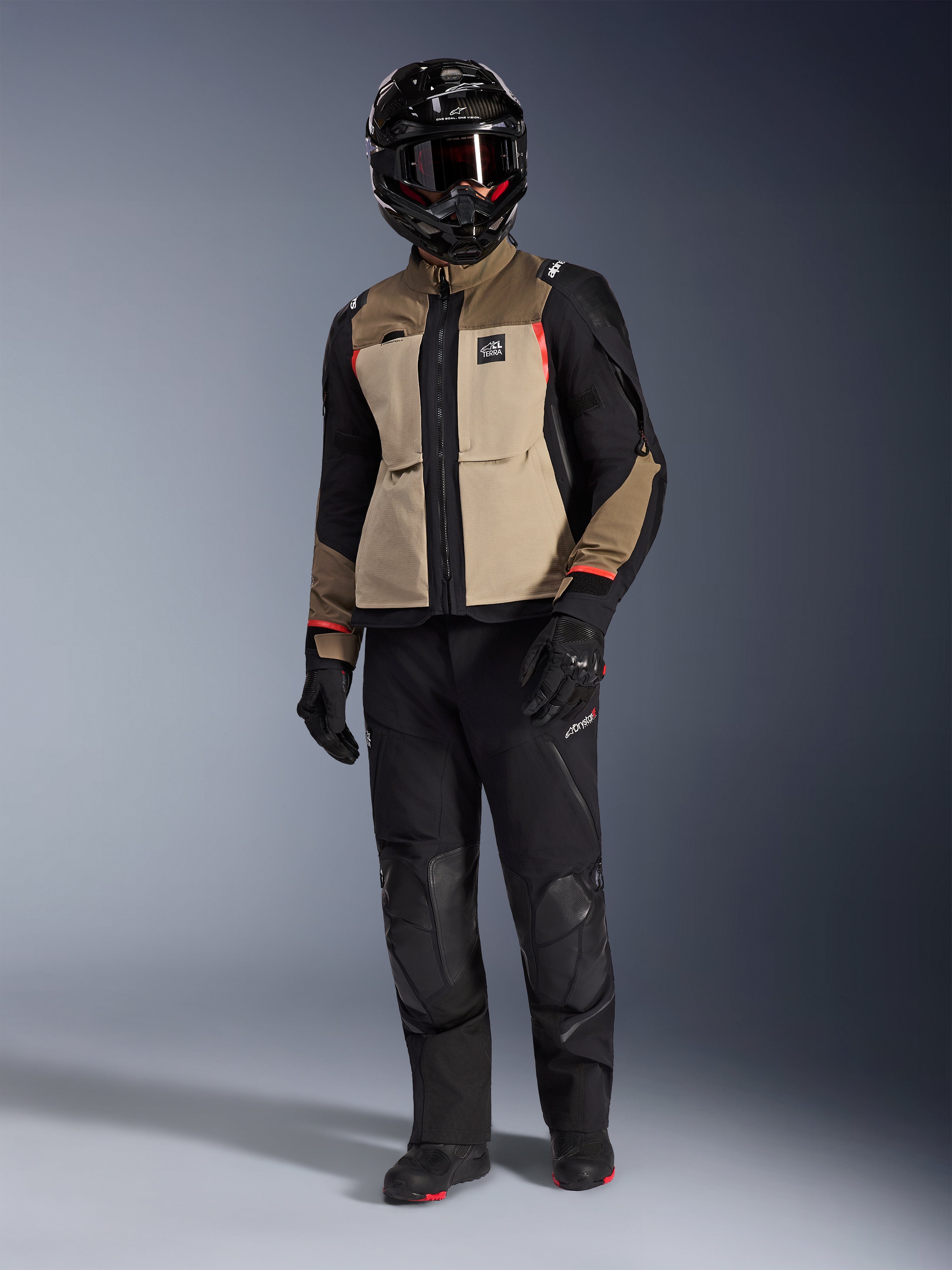 Alpinestars Andes Pro Drystar® XF-jakke i Walnut Aluminum, adventure-motorcykeljakke kombineret med matchende sorte bukser, en sort adventure-hjelm med briller og sorte støvler, med tan-farvede og sorte paneler med røde detaljer.