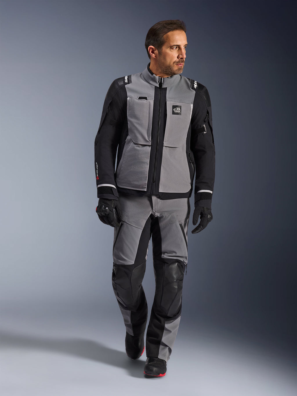 Mandlig model iført Alpinestars Andes Pro Drystar® XF-jakke i mørkegrå og sort, adventure motorcykel-køredragt med matchende bukser og handsker, med en teknisk tekstilskal med 'All Terra'-branding, komplet udstyr til langdistance adventure-touring