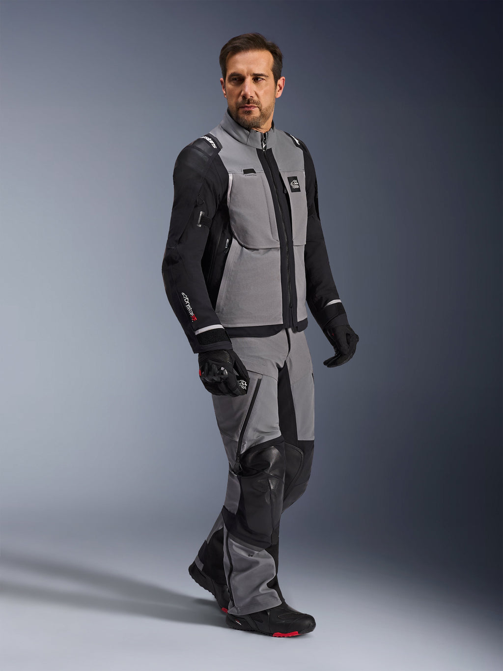 Alpinestars Andes Pro Drystar® XF Jakke, adventure motorcykeljakke, mørkegrå og sort, båret af en model med matchende tekstilbukser og sorte handsker, med design med flere lommer, teknisk 3-lags lamineret skal og artikuleret pasform til langdistance adventure-kørsel.