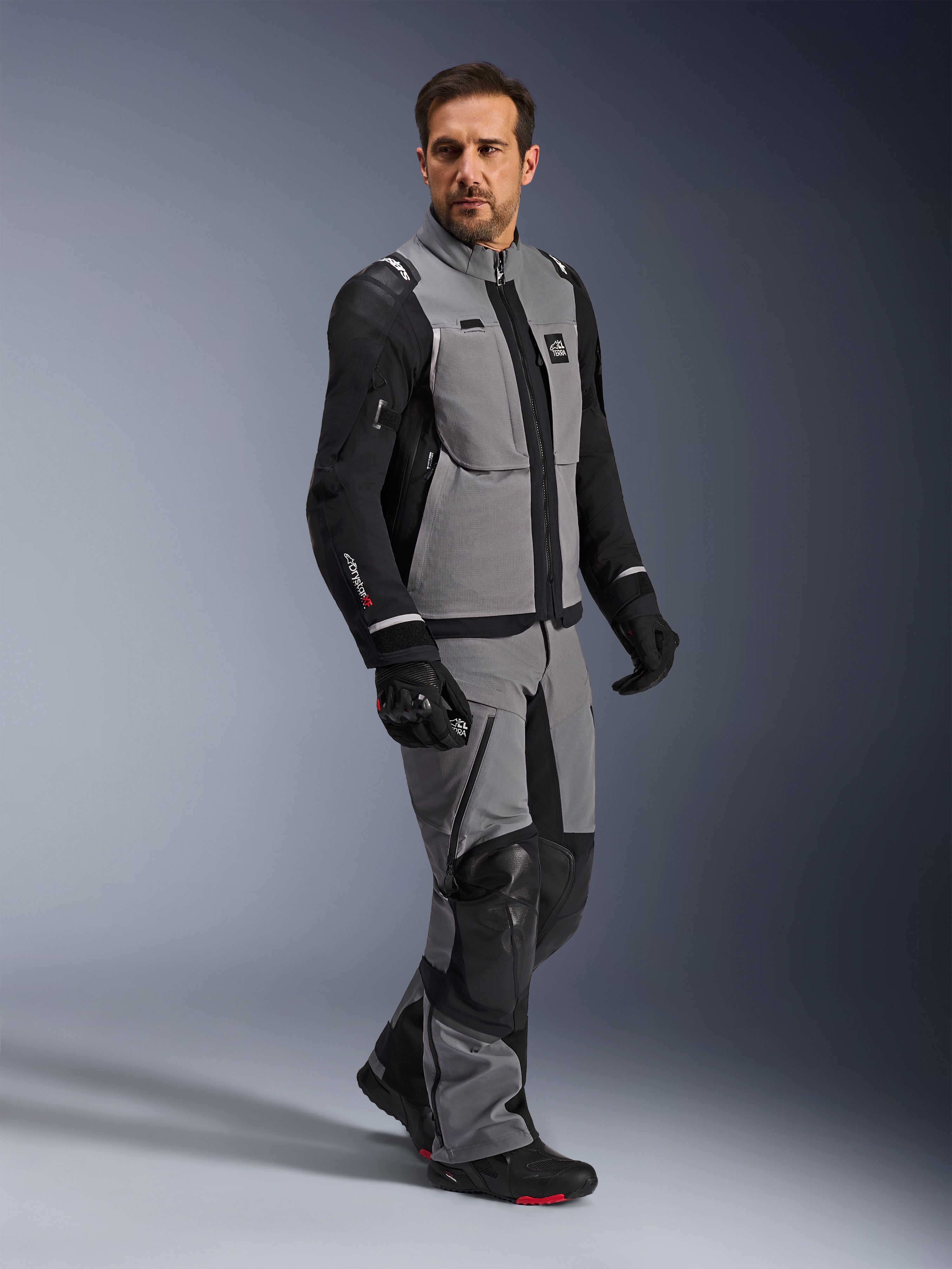 Alpinestars Andes Pro Drystar® XF Jakke, adventure motorcykeljakke, mørkegrå og sort, båret af en model med matchende tekstilbukser og sorte handsker, med design med flere lommer, teknisk 3-lags lamineret skal og artikuleret pasform til langdistance adventure-kørsel.