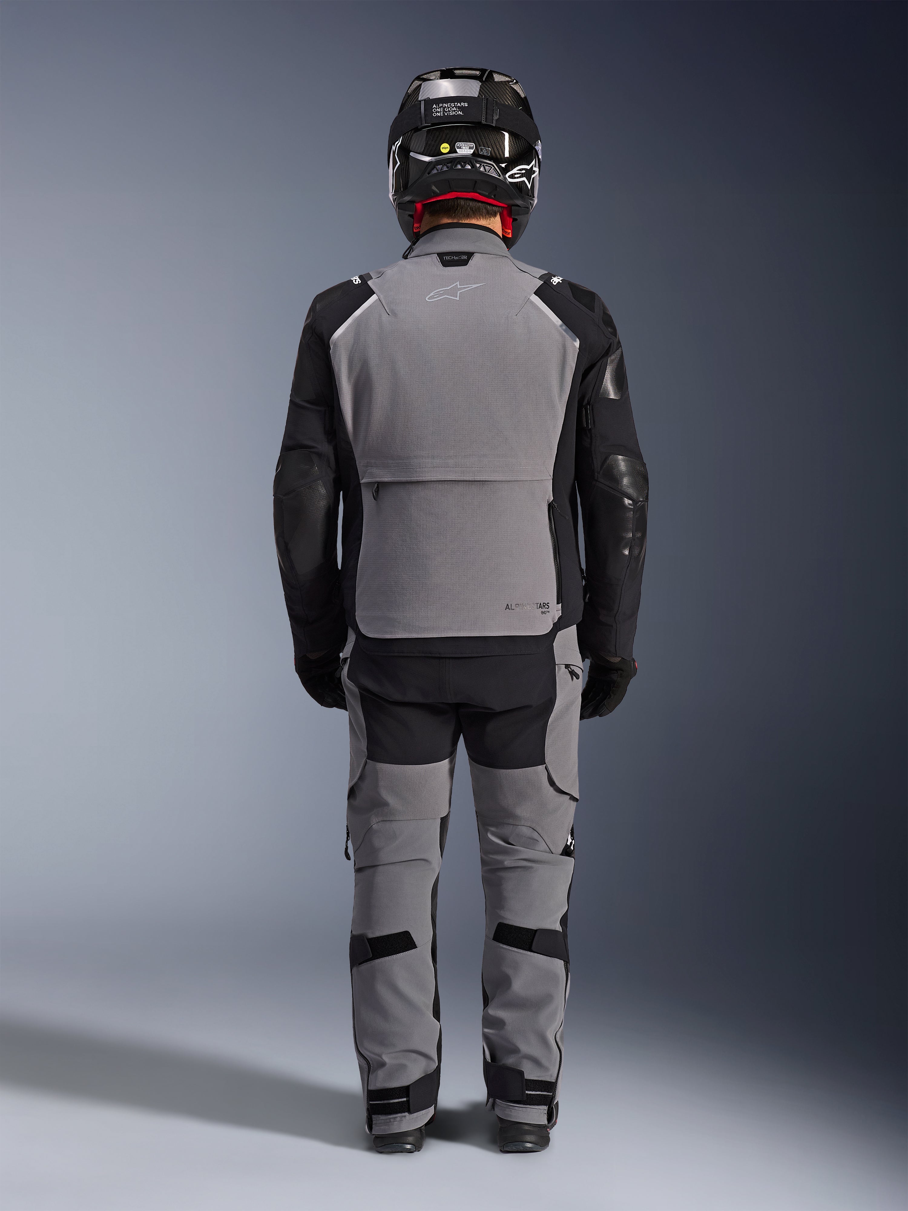 Alpinestars Andes Pro Drystar® XF-jakke, set bagfra, mørkegråt og sort adventure touring-udstyr, kombineret med matchende grå og sorte bukser, sort kulfiberhjelm og sorte handsker, med tekniske paneler og reflekterende branding