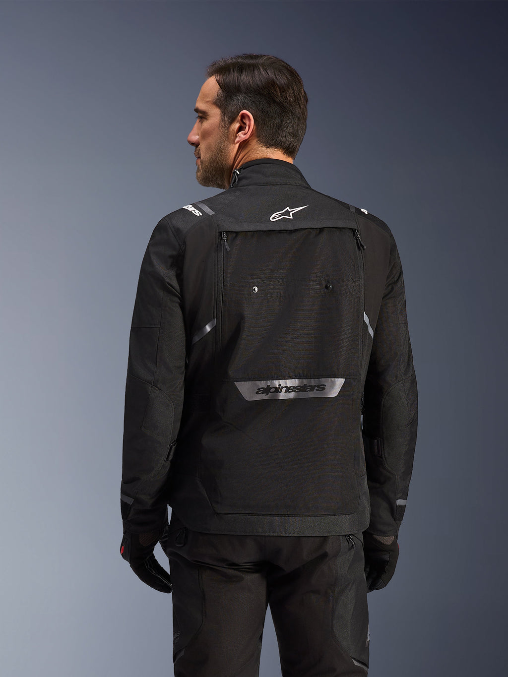 Motorcyklist set bagfra iført Alpinestars Cusco Drystar®-jakke i sort, tekstil adventure touring-jakke med stor opbevaringslomme bagpå og reflekterende branding, kombineret med matchende sorte bukser og handsker.