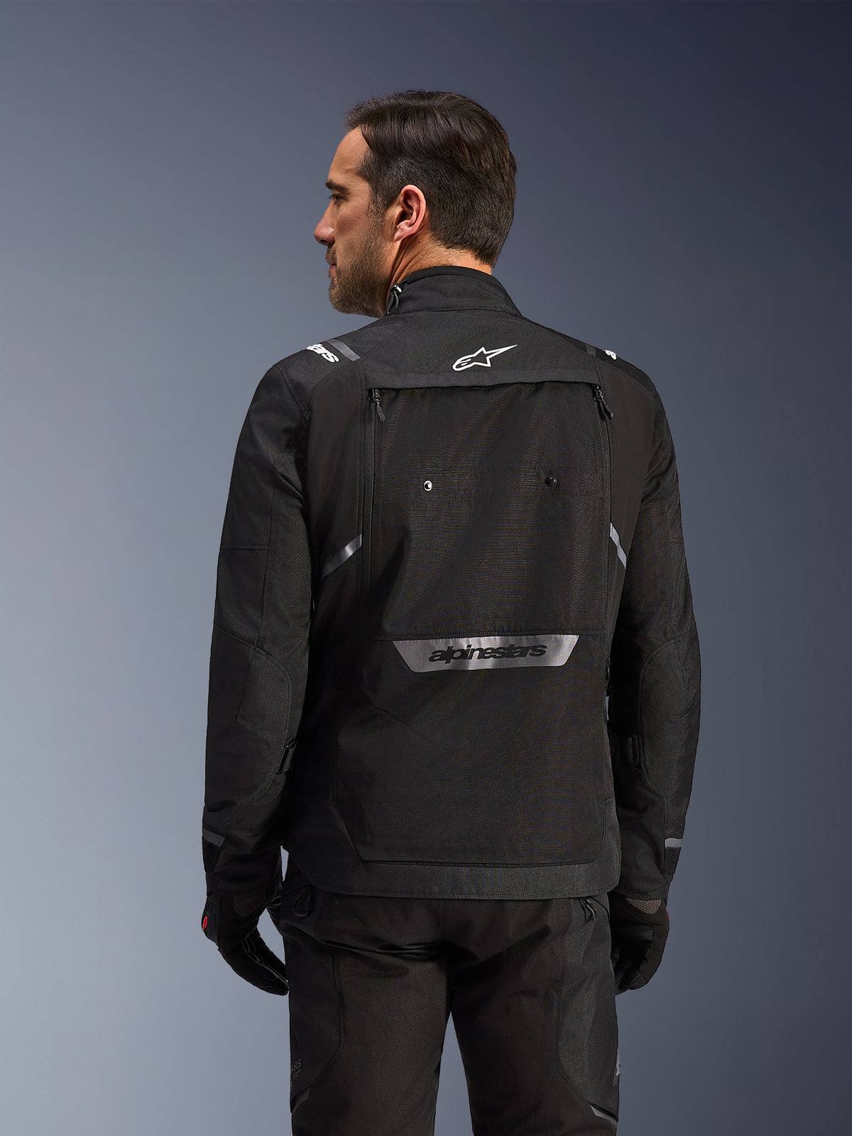Motorcyklist set bagfra iført Alpinestars Cusco Drystar®-jakke i sort, tekstil adventure touring-jakke med stor opbevaringslomme bagpå og reflekterende branding, kombineret med matchende sorte bukser og handsker.