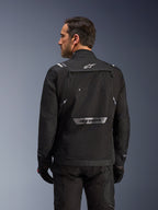 Motorcyklist set bagfra iført Alpinestars Cusco Drystar®-jakke i sort, tekstil adventure touring-jakke med stor opbevaringslomme bagpå og reflekterende branding, kombineret med matchende sorte bukser og handsker.