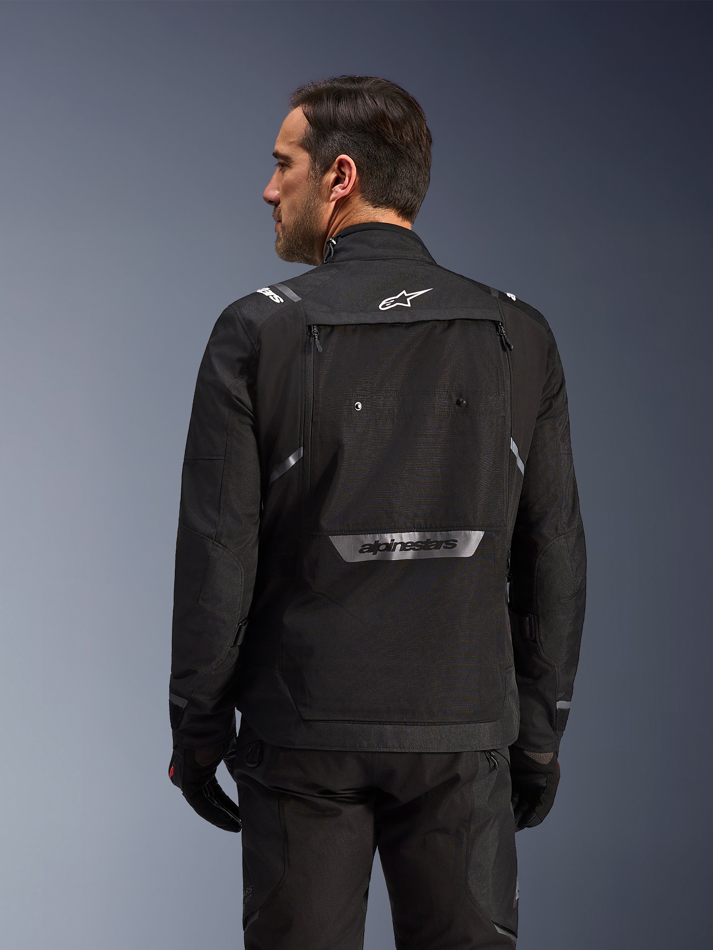 Motorcyklist set bagfra iført Alpinestars Cusco Drystar®-jakke i sort, tekstil adventure touring-jakke med stor opbevaringslomme bagpå og reflekterende branding, kombineret med matchende sorte bukser og handsker.