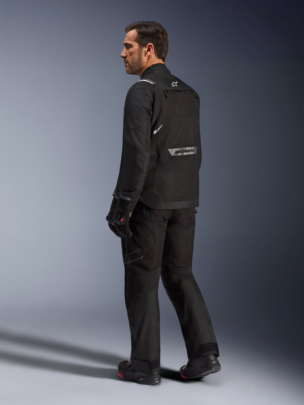 Alpinestars Cusco Drystar® jakke, sort motorcykeljakke, set bagfra på mandlig model i fuldt køreudstyr, med reflekterende branding på ryggen, tekstilkonstruktion med tekniske paneler, koordineret sæt til road adventure touring