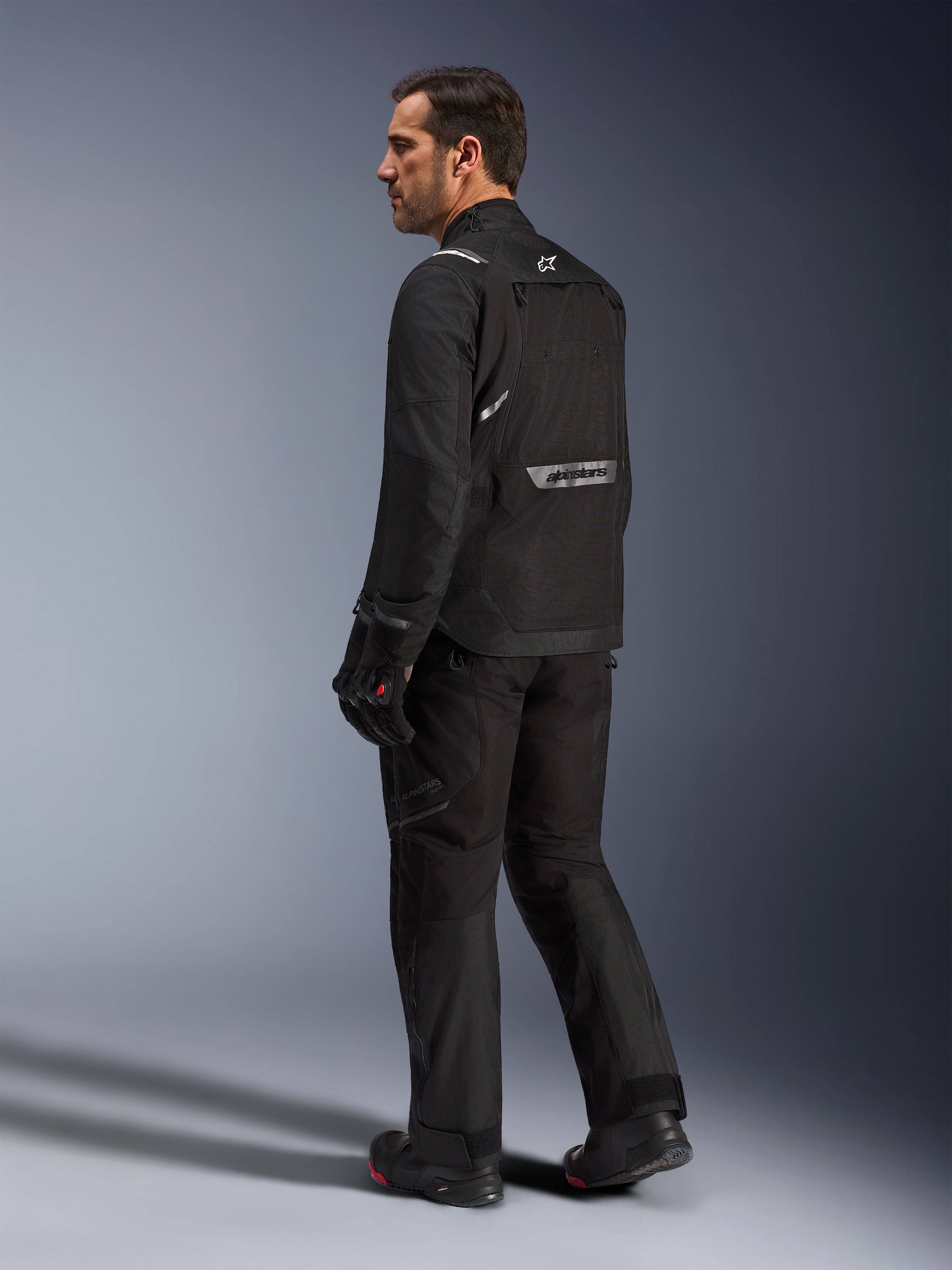 Alpinestars Cusco Drystar® jakke, sort motorcykeljakke, set bagfra på mandlig model i fuldt køreudstyr, med reflekterende branding på ryggen, tekstilkonstruktion med tekniske paneler, koordineret sæt til road adventure touring