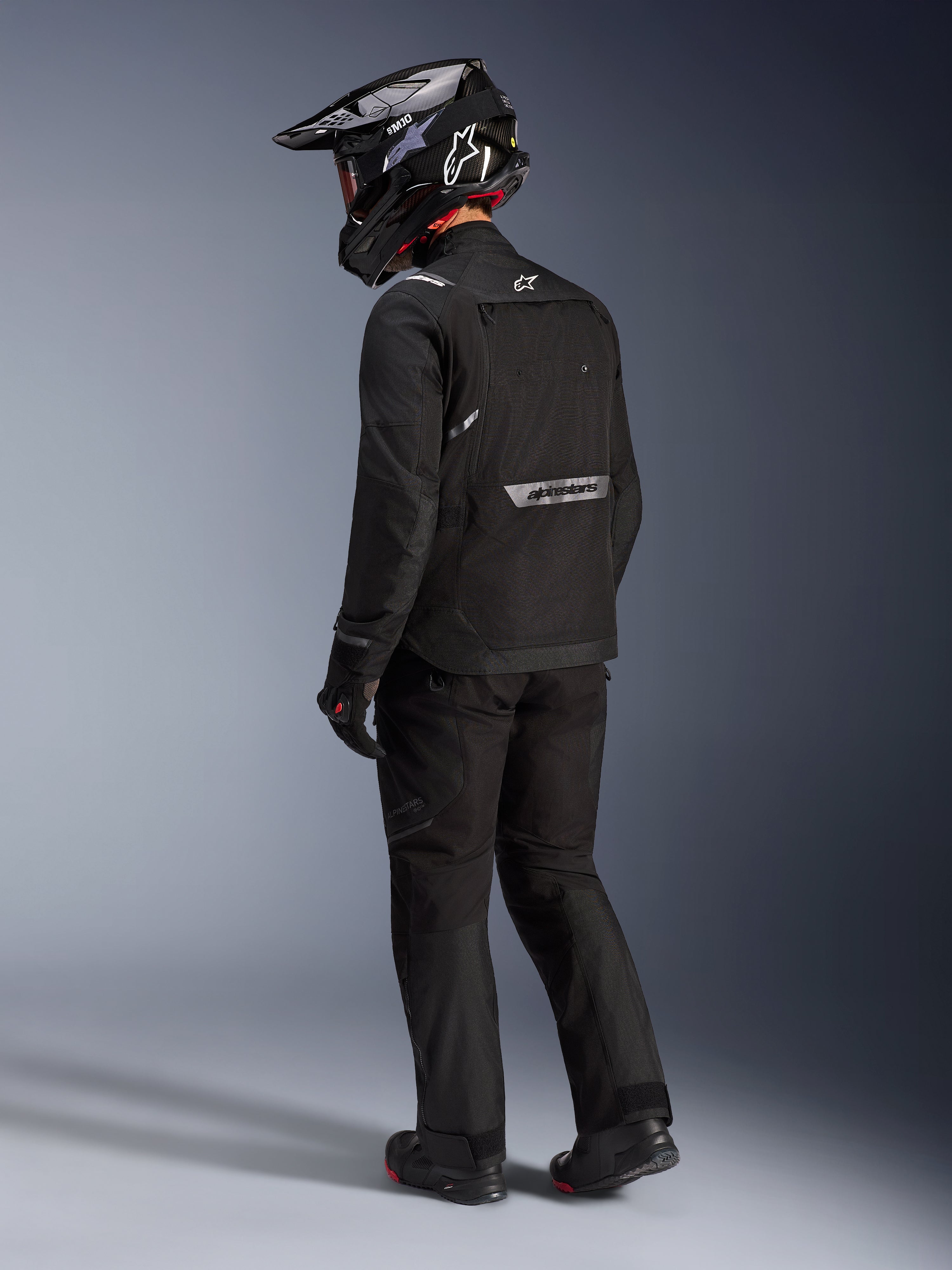 Alpinestars Cusco Drystar®-jakke i sort, set bagfra fra siden af en kører iført et komplet adventure touring-sæt, inklusive matchende sorte bukser og handsker, sort adventure-hjelm, tekstilkonstruktion med reflekterende branding på ryggen