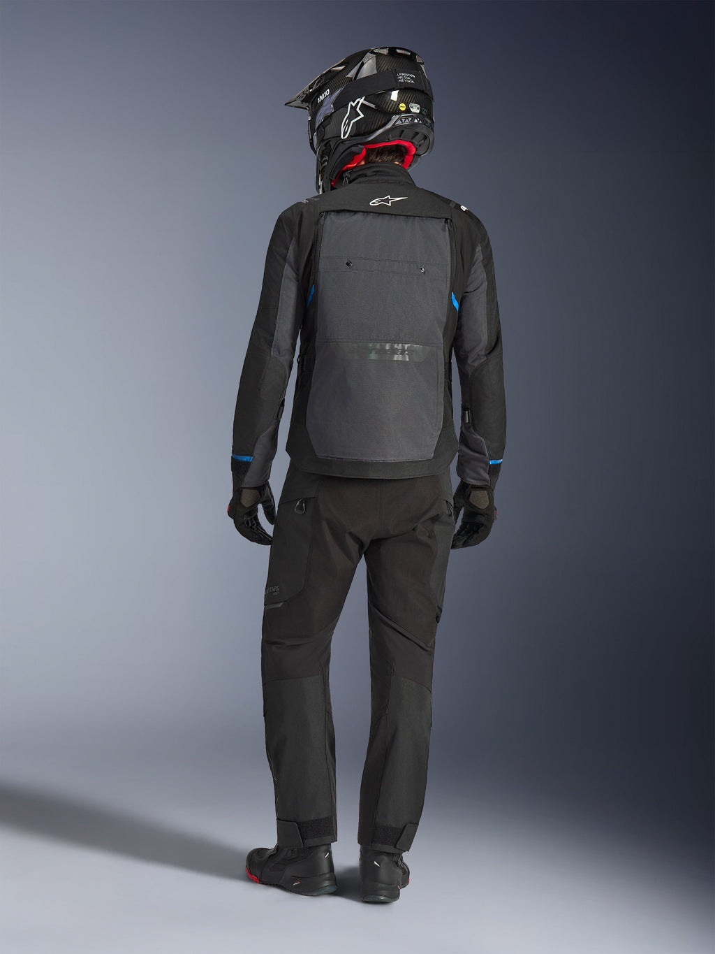 Alpinestars Cusco Drystar® jakke, sort og mørkegrå, set bagfra af en kører iført komplet adventure touring-udstyr, stort gråt rygpanel med Alpinestars-logo, blå detaljer på ærmerne, båret med matchende sorte bukser og hjelm.