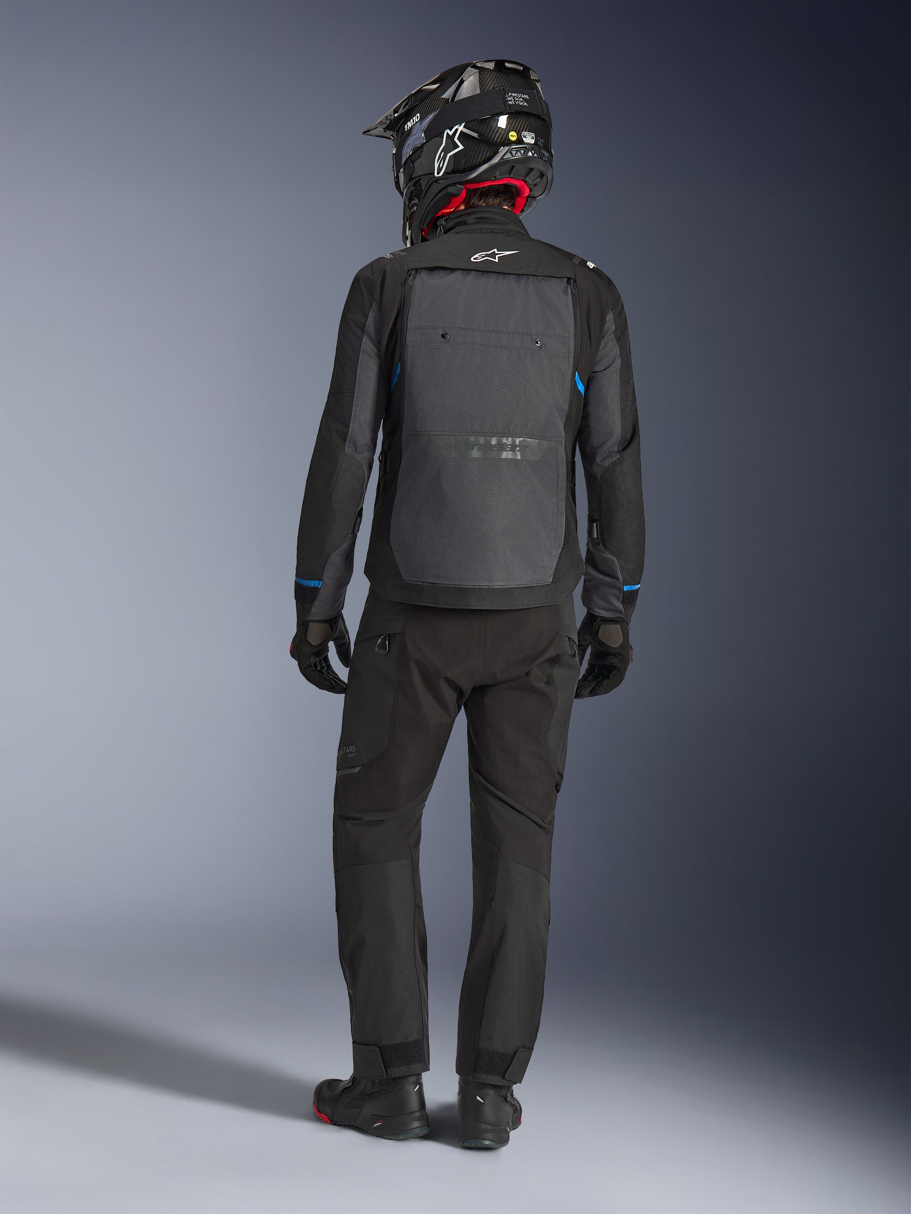 Alpinestars Cusco Drystar® jakke, sort og mørkegrå, set bagfra af en kører iført komplet adventure touring-udstyr, stort gråt rygpanel med Alpinestars-logo, blå detaljer på ærmerne, båret med matchende sorte bukser og hjelm.