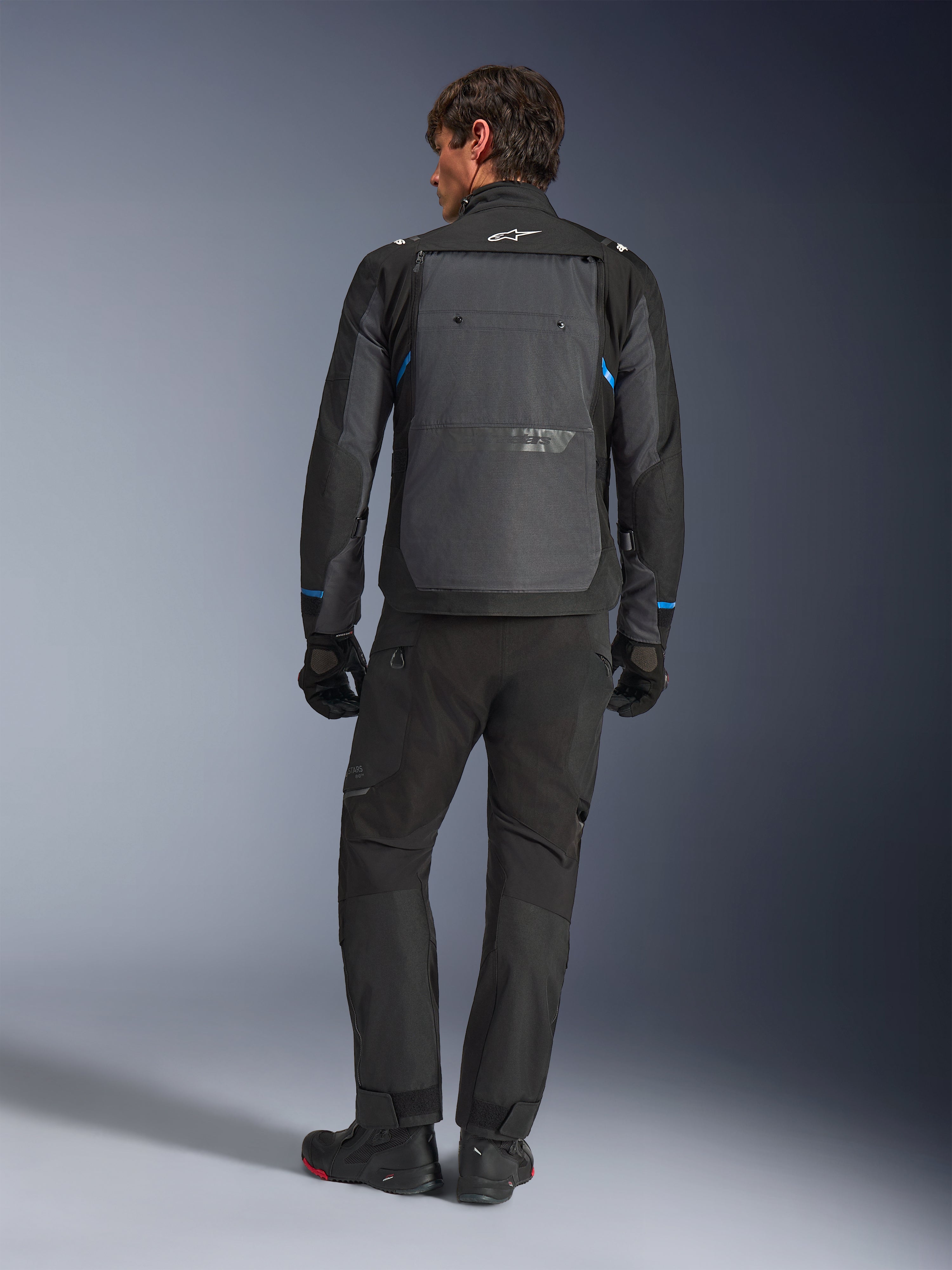Alpinestars Cusco Drystar® Jacket, sort og mørkegrå med blå detaljer, set bagfra, båret med matchende sorte adventure-bukser og handsker, med et stort gråt rygpanel og hvidt logo på kraven, designet til vej- og adventure-touring