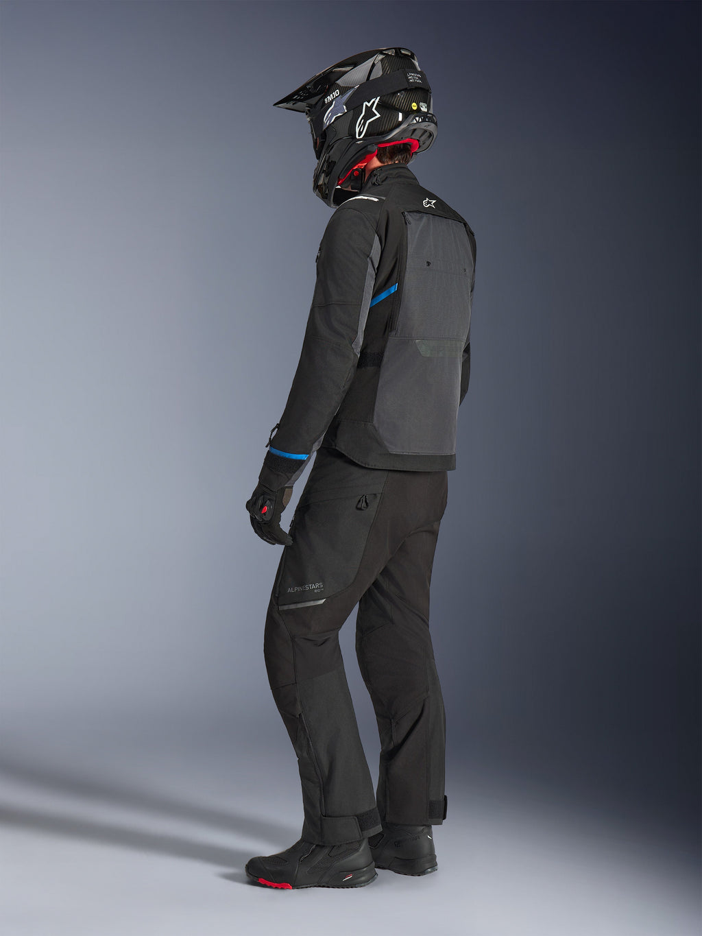 Motorcykelrytter iført Alpinestars Cusco Drystar® jakke i sort og mørkegrå med blå detaljer, kombineret med sorte kørebukser, en SM10-hjelm i kulfiber og sorte handsker, set bagfra med det store ventilationspanel og adventure touring-design.