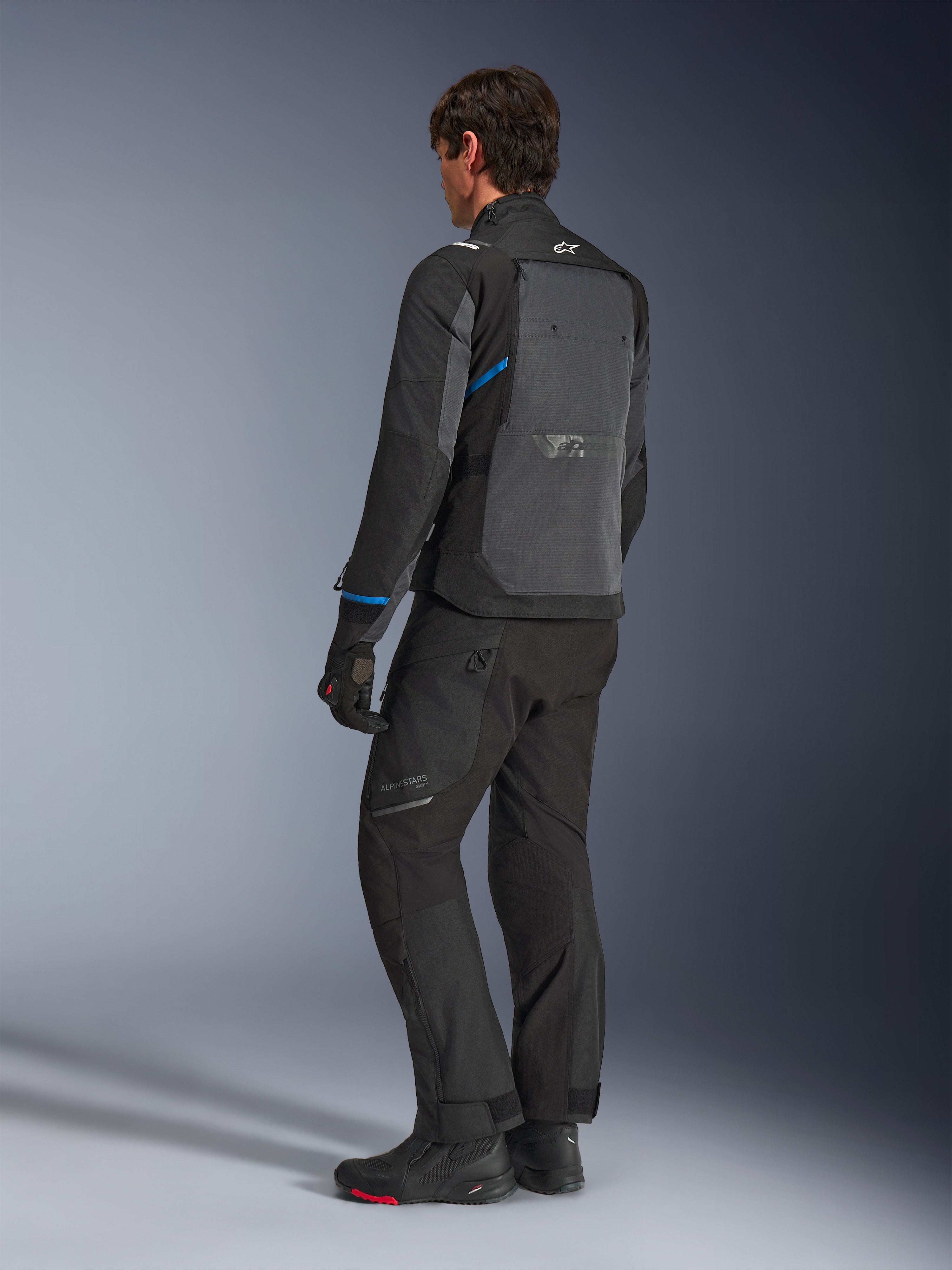 Alpinestars Cusco Drystar® jakke, sort og mørkegrå, set bagfra i trekvart profil, der viser et stort ventilationspanel på ryggen og blå detaljer på ærmerne, båret med sorte motorcykelbukser og støvler for et komplet touring-outfit.