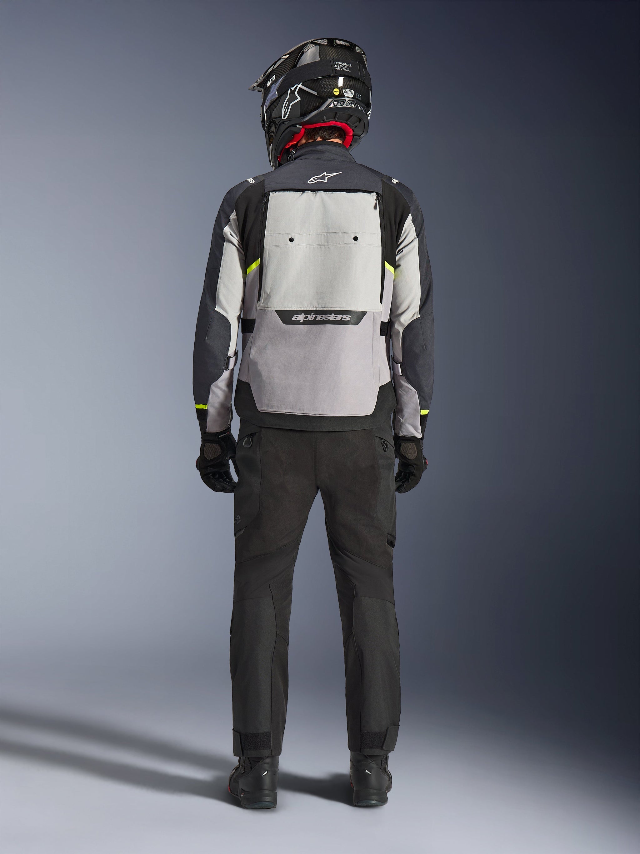 Motorcycle rider seen from behind wearing the Alpinestars Cusco Drystar® Jacket in Ice Gray, Mid Gray, and Deep Gray, featuring a large back ventilation panel and Alpinestars branding, paired with black riding pants and a black helmet, adventure touring motorcycle apparel