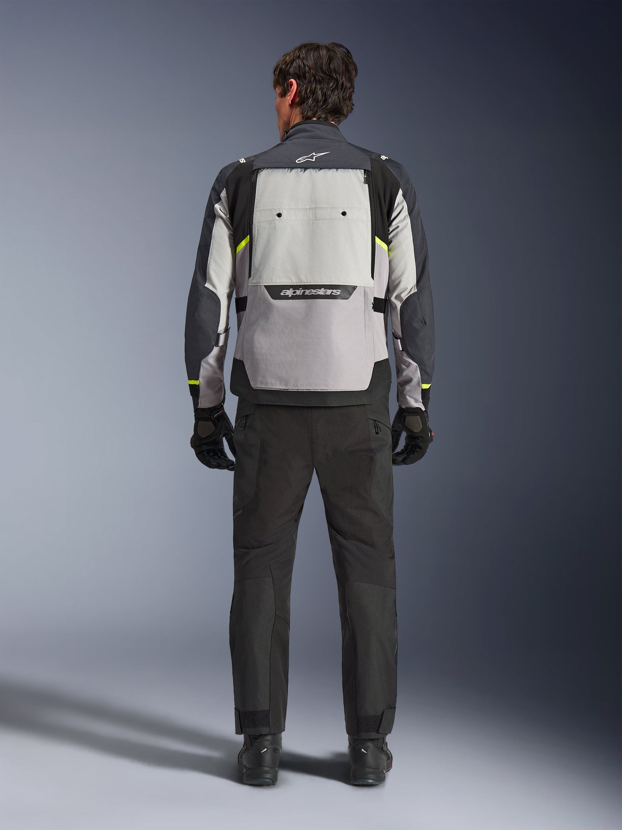 Rear view of a rider wearing the Alpinestars Cusco Drystar® Jacket in Ice Gray, Mid Gray, and Deep Gray, styled with black motorcycle pants and gloves, highlighting the large rear ventilation panel and Alpinestars branding