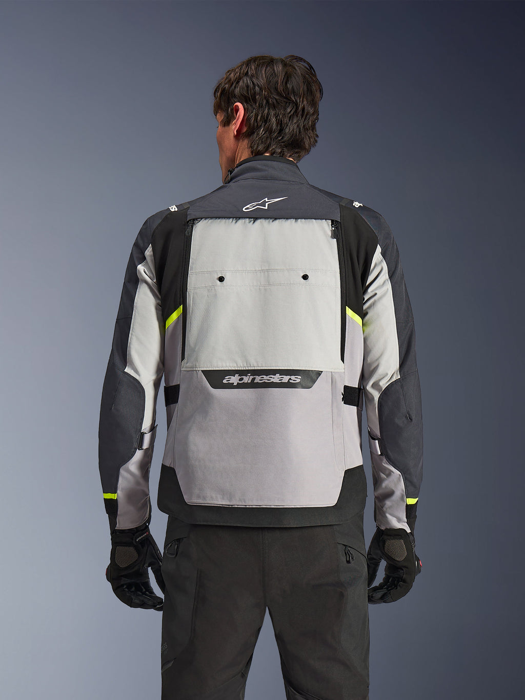 Set bagfra af en kører iført Alpinestars Cusco Drystar®-jakken i Ice Gray, Mid Gray og Deep Gray, med neongule detaljer, et stort ventilationspanel på ryggen og markant Alpinestars-branding, kombineret med sorte kørebukser og handsker.
