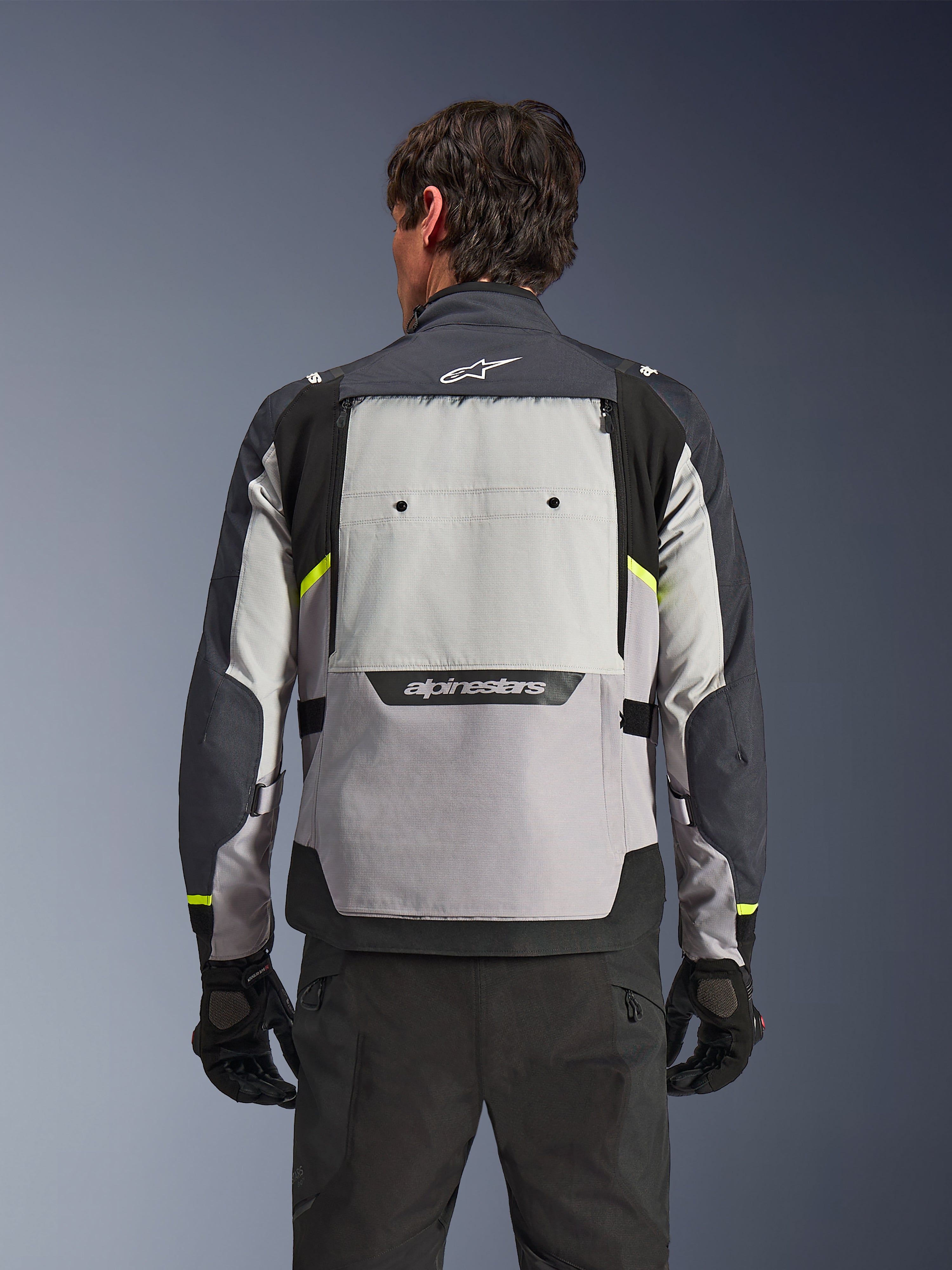 Set bagfra af en kører iført Alpinestars Cusco Drystar®-jakken i Ice Gray, Mid Gray og Deep Gray, med neongule detaljer, et stort ventilationspanel på ryggen og markant Alpinestars-branding, kombineret med sorte kørebukser og handsker.