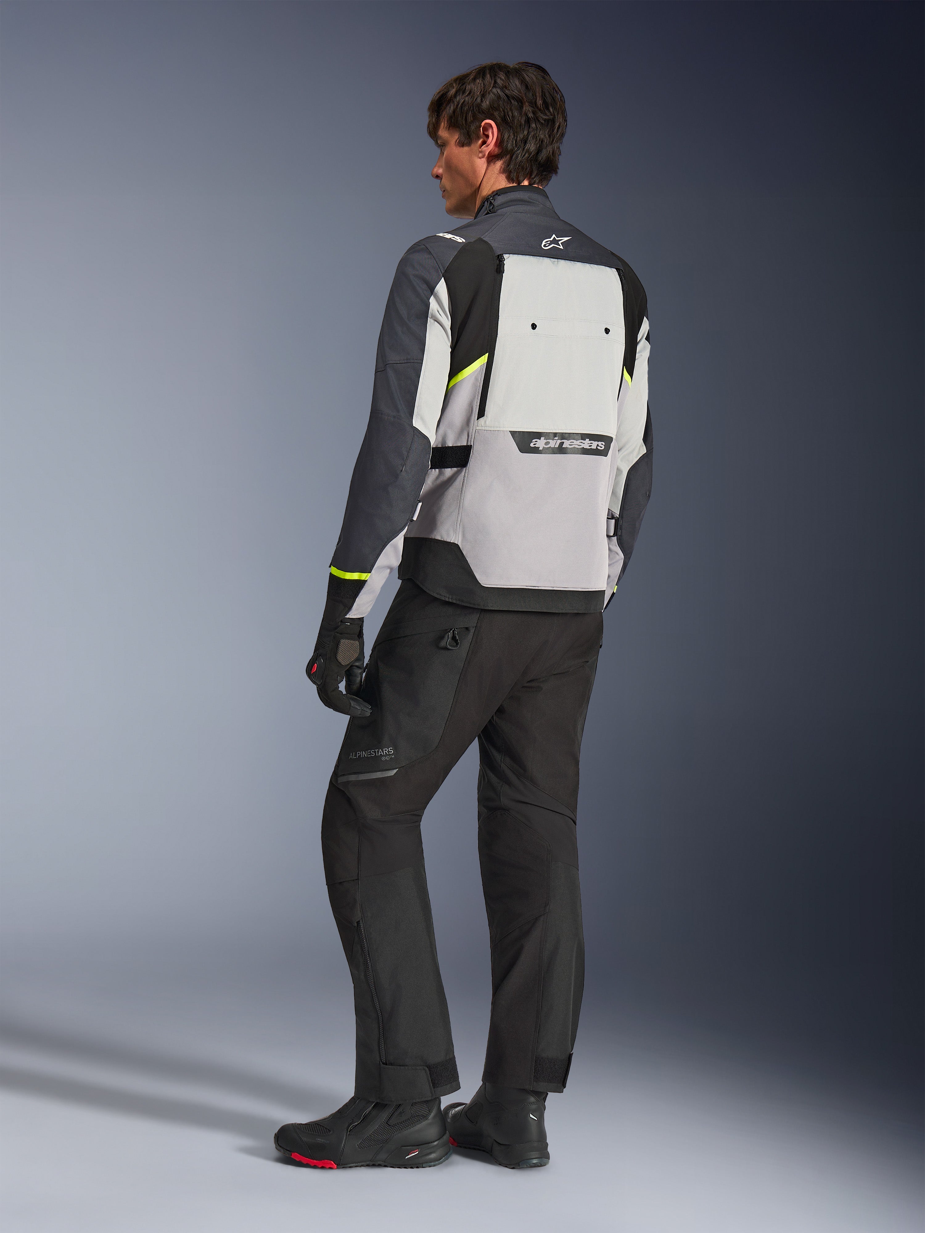 Alpinestars Cusco Drystar®-jakke i Ice Gray, Mid Gray og Deep Gray, set bagfra på model, adventure touring-jakke med neongule detaljer, båret med sorte kørebukser og støvler, med et stort ventilationspanel på ryggen og Alpinestars-branding.