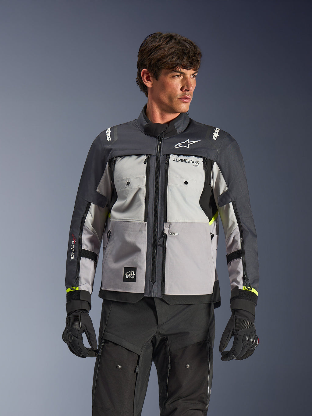 Alpinestars Cusco Drystar® Jakke, adventure touring motorcykeljakke, Ice Gray, Mid Gray og Deep Gray, båret af en model med sorte bukser og handsker, teknisk tekstildesign med flere paneler, brystlommer, 'Drystar'-branding på ærmet og subtile neongule detaljer ved manchetterne.