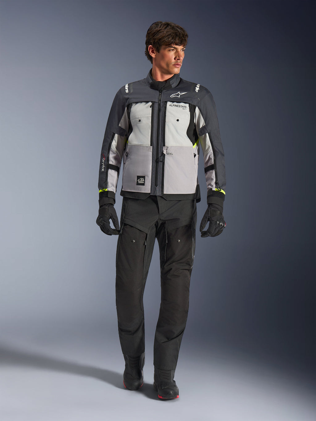 Alpinestars Cusco Drystar®-jakke i Ice Gray, Mid Gray og Deep Gray, båret med sorte adventure-motorcykelbukser og -handsker, tekstil adventure-touringjakke med grå paneler i flere nuancer og neongule detaljer, studiefoto af mandlig model.