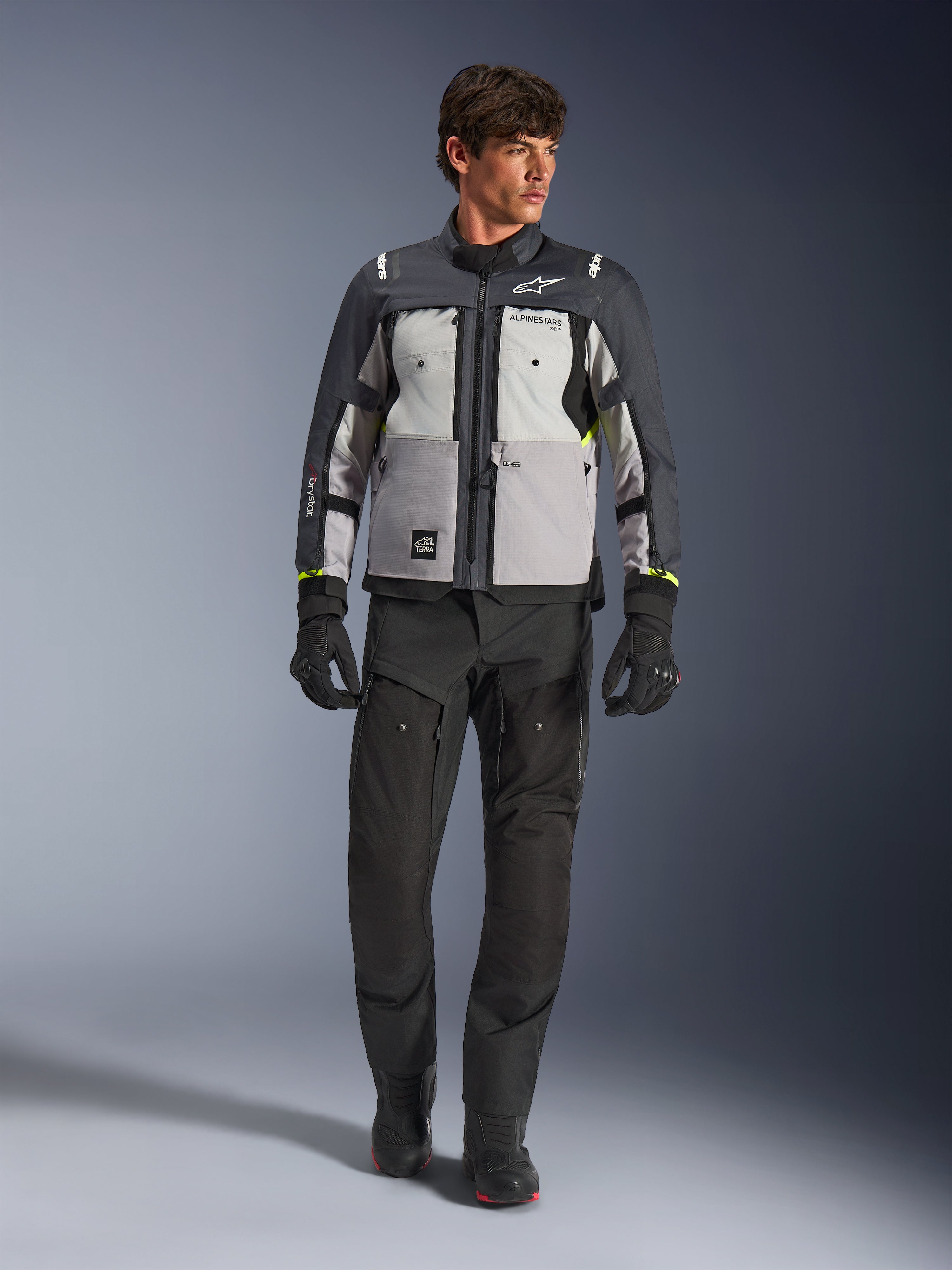 Alpinestars Cusco Drystar®-jakke i Ice Gray, Mid Gray og Deep Gray, båret med sorte adventure-motorcykelbukser og -handsker, tekstil adventure-touringjakke med grå paneler i flere nuancer og neongule detaljer, studiefoto af mandlig model.