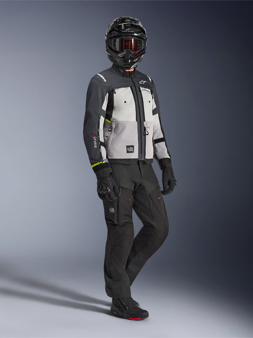 Alpinestars Cusco Drystar® jakke i Ice Gray Mid Gray Deep Gray, fuldkropsbillede af en kører i komplet adventure-udstyr, inklusiv sort hjelm med røde goggles, matchende sorte bukser og handsker, tekstil-motorcykeljakke med direkte ventilationspaneler og vandtæt membran
