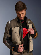 Alpinestars Cusco Drystar® jakke, Walnut Aluminum Black, mand der demonstrerer brystbeskytterlommen med en rød beskyttelsesindsats, tekstil motorcykeljakke med ventilationsåbninger med lynlås, branding til adventure touring