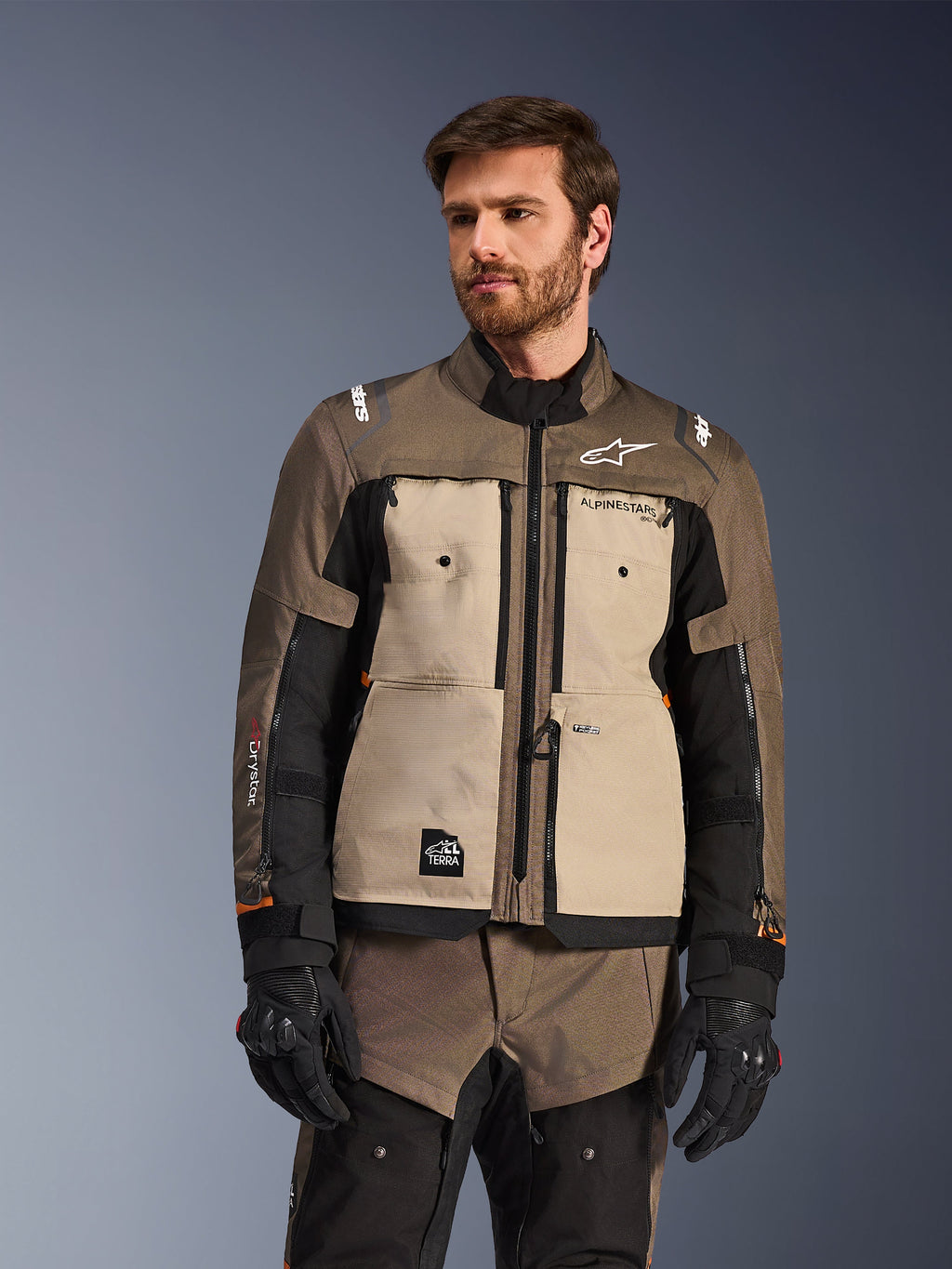 Alpinestars Cusco Drystar® jakke, adventure motorcykeljakke, Walnut Aluminum Black, vist på en model med sorte handsker, med sandfarvede brystpaneler og sorte ærmer med Alpinestars-branding og All Terra-mærke, beskyttende touring-udstyr til al slags vejr