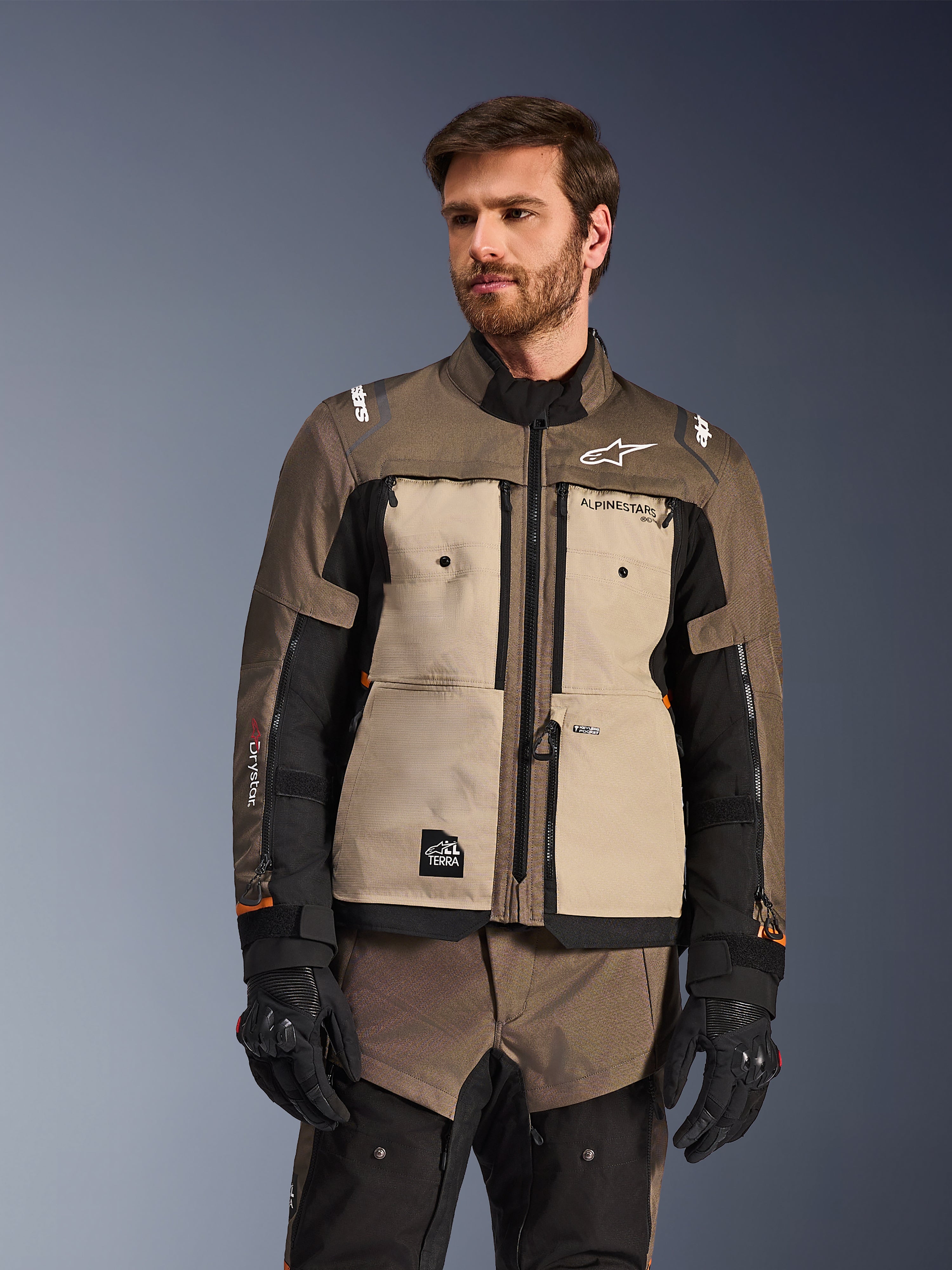 Alpinestars Cusco Drystar® jakke, adventure motorcykeljakke, Walnut Aluminum Black, vist på en model med sorte handsker, med sandfarvede brystpaneler og sorte ærmer med Alpinestars-branding og All Terra-mærke, beskyttende touring-udstyr til al slags vejr