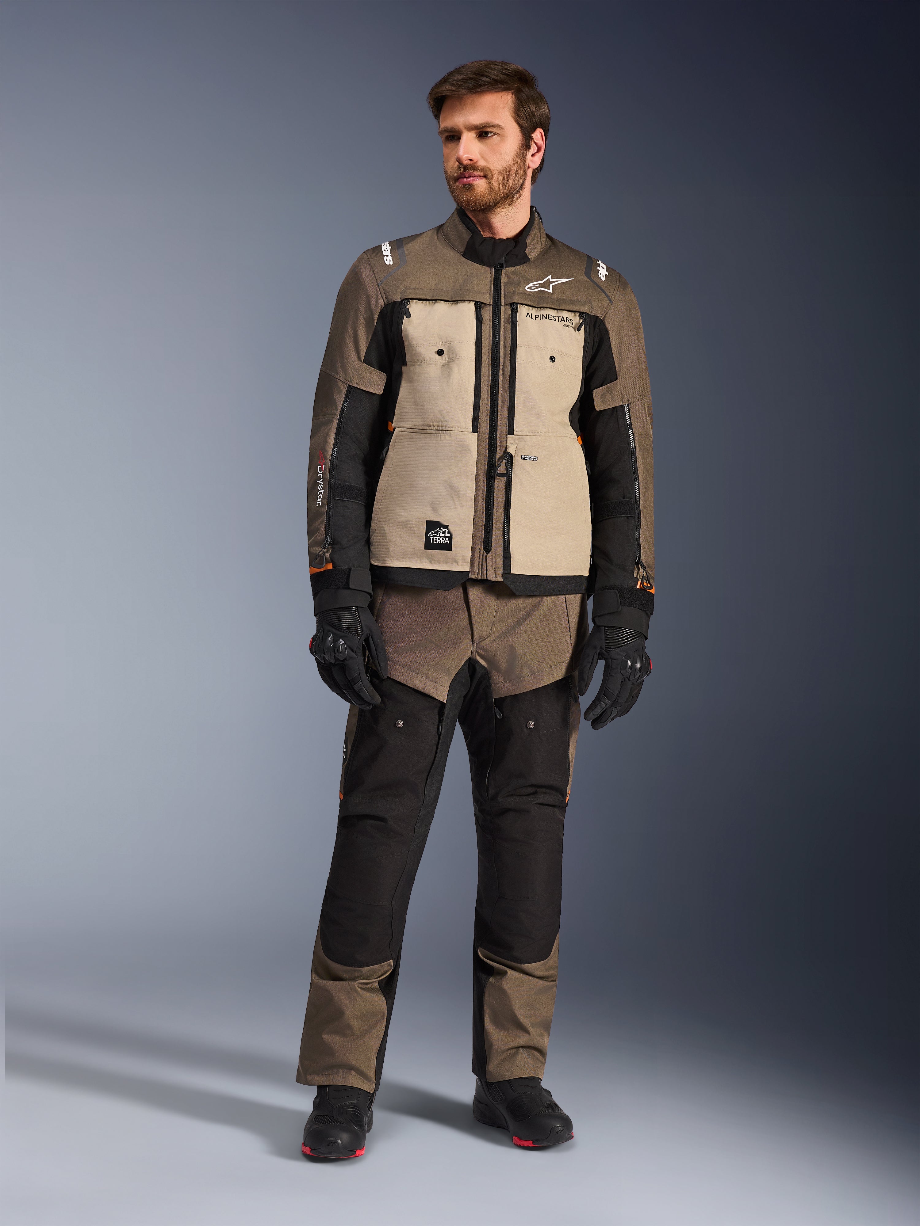 Alpinestars Cusco Drystar®-jakke i Walnut Aluminum Black, båret af en mandlig kører med matchende adventure touring-bukser, sorte handsker og støvler, med sandfarvede brystpaneler, sorte ærmer med diskrete orange detaljer og funktionelle ventilationslynlåse til motorcykelkørsel på vej.