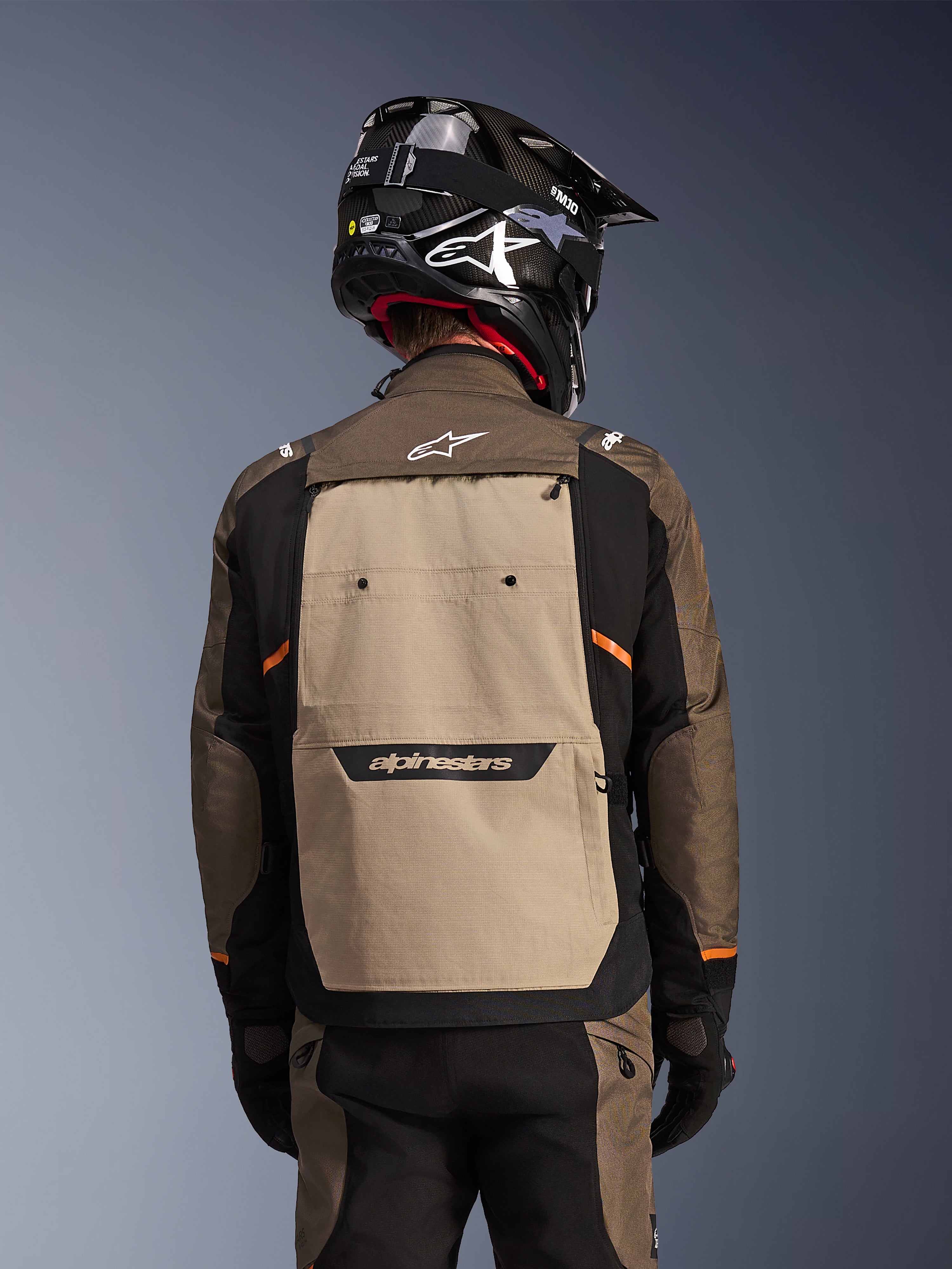 Set bagfra af Alpinestars Cusco Drystar®-jakken i Walnut Aluminum Black, adventure touring motorcykeljakke med et stort sandfarvet rygpanel med Alpinestars-branding, sorte ærmer med orange detaljer, vist med en sort hjelm i carbon-stil.