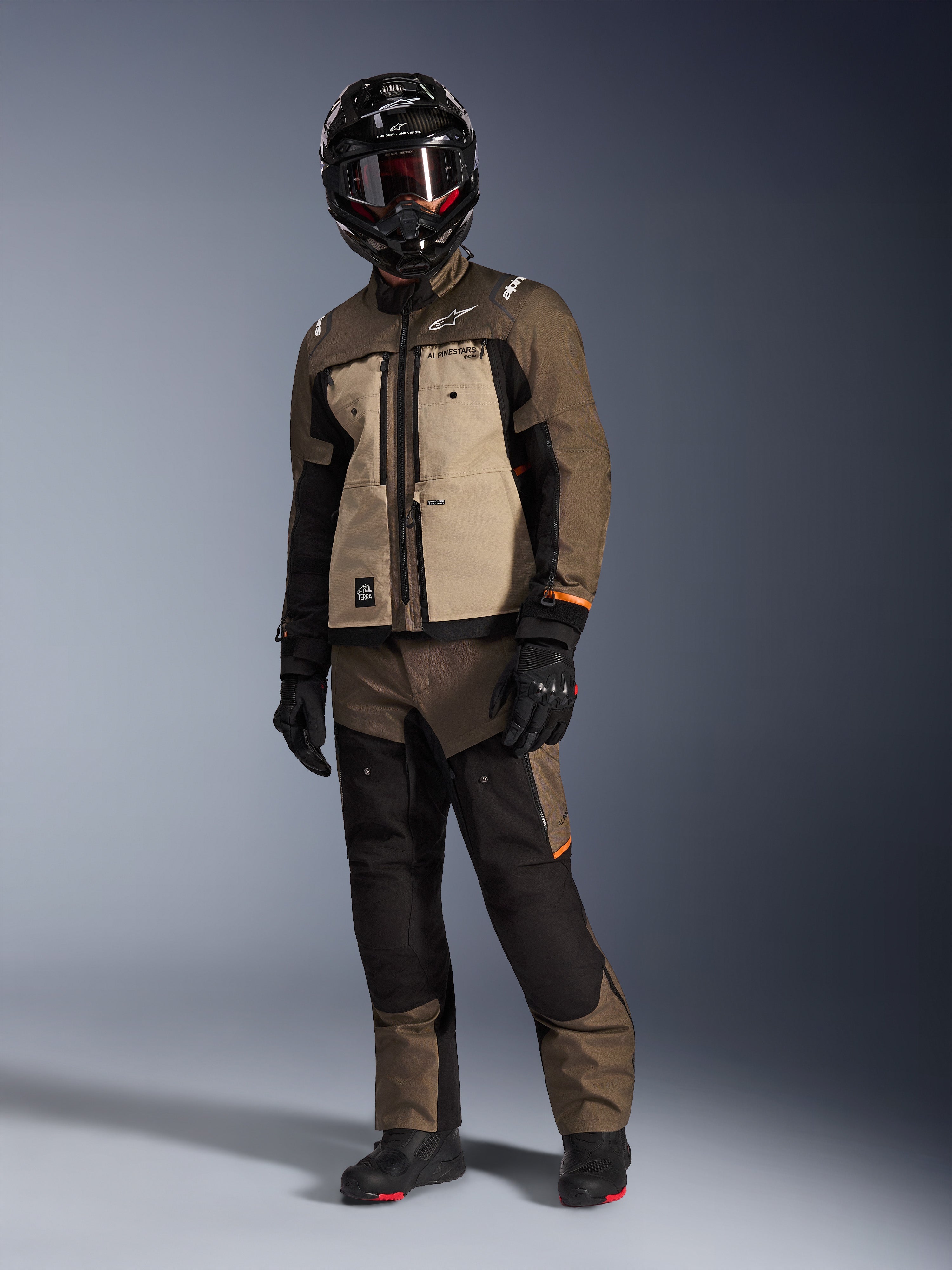 Alpinestars Cusco Drystar®-jakke, Walnut Aluminum Black, båret af en kører i komplet adventure touring-udstyr, med beige og brune paneler og sorte detaljer, koordineret med matchende bukser, en sort full-face hjelm med goggles og sorte støvler, studiobaggrund