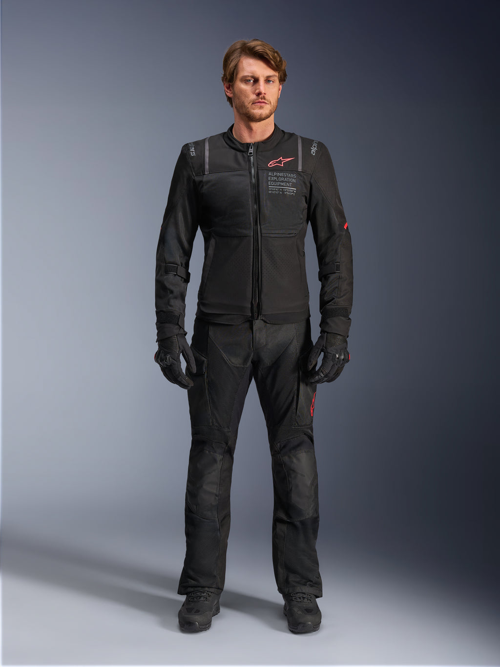 Mandlig model iført Alpinestars ST-2 Air-jakke, sort motorcykeljakke med mesh-paneler og rødt logo, 'Alpinestars Exploration Equipment'-tekst på brystet, kombineret med sorte tekniske kørebukser og handsker, moderne adventure touring-udstyr