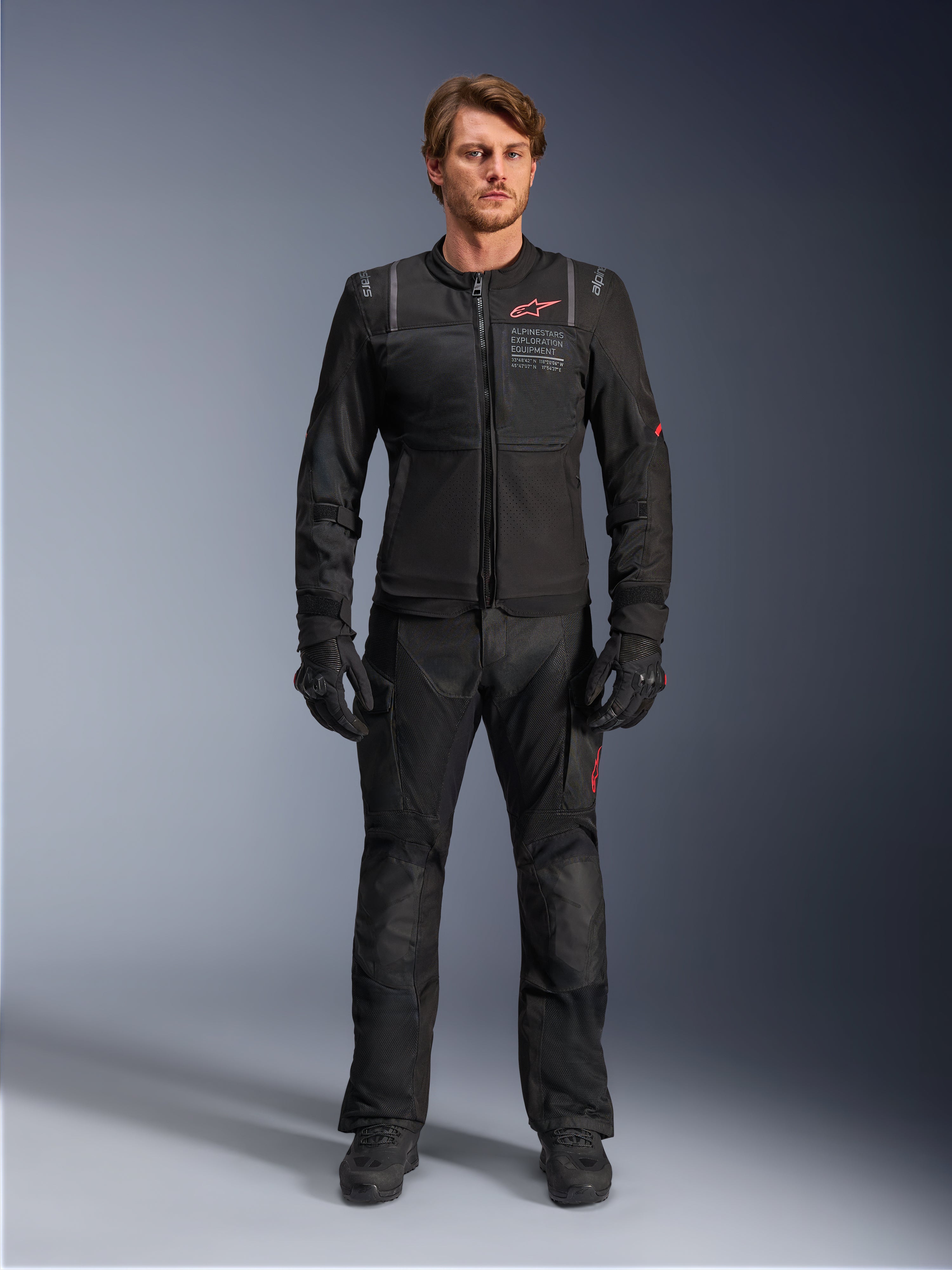 Mandlig model iført Alpinestars ST-2 Air-jakke, sort motorcykeljakke med mesh-paneler og rødt logo, 'Alpinestars Exploration Equipment'-tekst på brystet, kombineret med sorte tekniske kørebukser og handsker, moderne adventure touring-udstyr