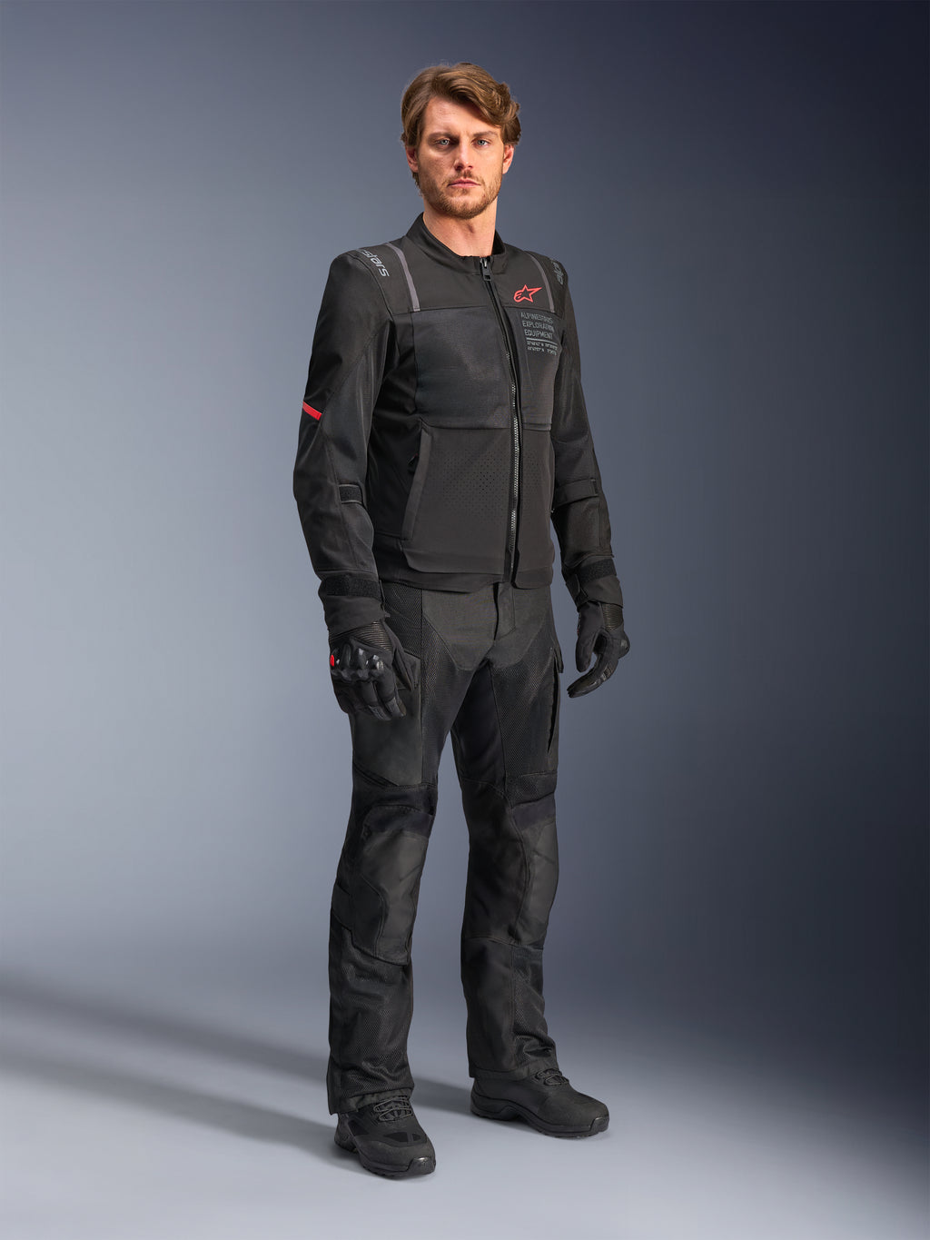 Mandlig model iført Alpinestars ST-2 Air-jakken i sort, med et rødt stjernelogo og 'Alpinestars Exploration Equipment'-branding, kombineret med matchende sorte kørebukser, handsker og støvler til et moderne sommer-touring-outfit.