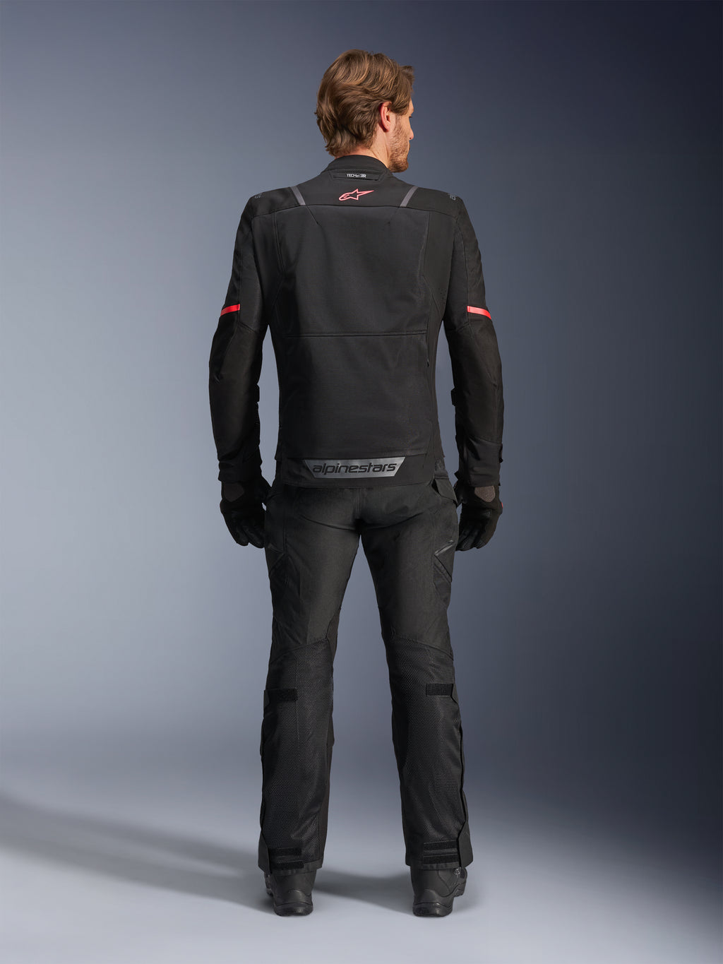 Alpinestars ST-2 Air jakke, sort, set bagfra af en kører iført motorcykeljakken og matchende sorte bukser, med mesh-ventilationspaneler, rødt logo øverst på ryggen og grå Alpinestars-branding på den nederste rygkant